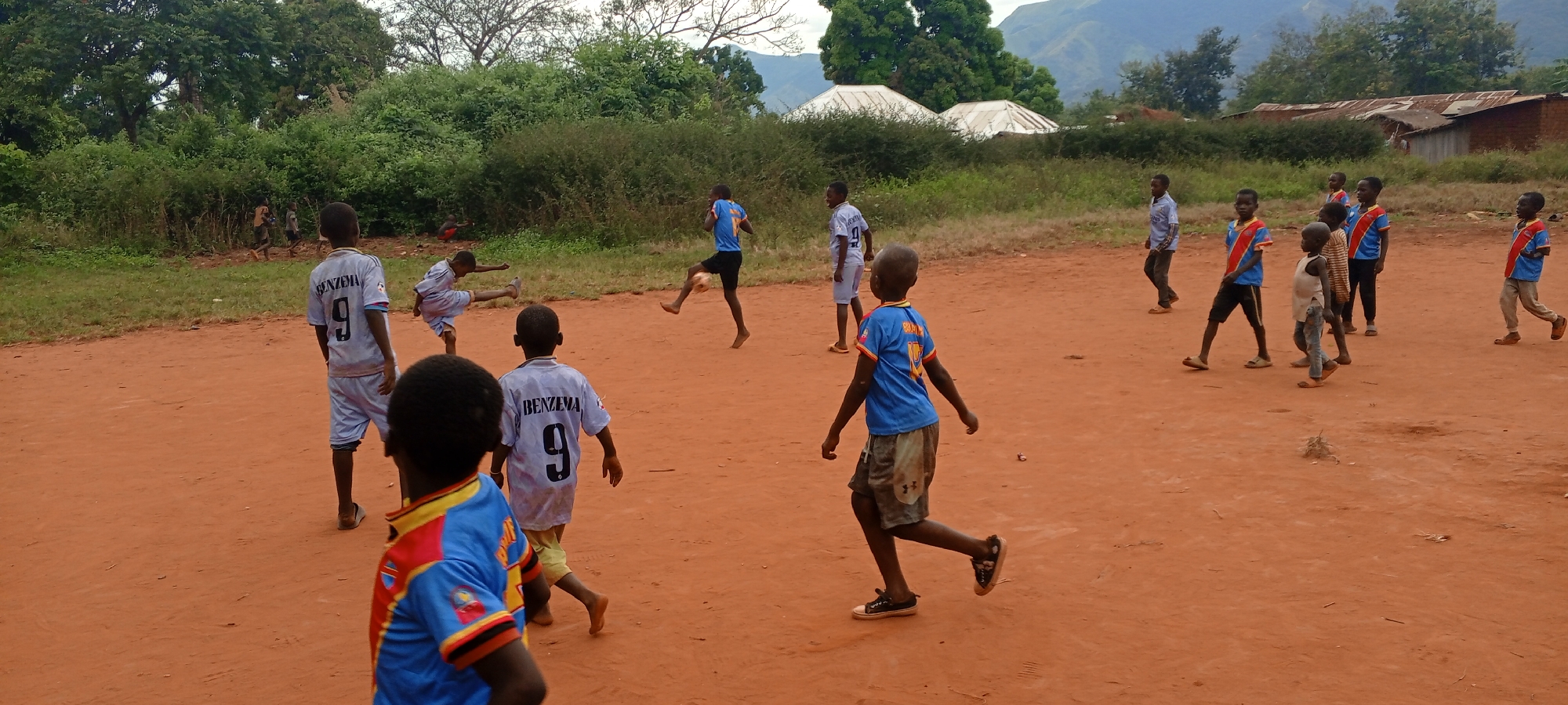 In Ngalula, Democratic Republic of the Congo, a group of children plays on a reddish-brown dirt field. Most of the children are barefoot, while a few wear sandals or shoes. Some of them are wearing soccer jerseys, including blue ones with a red and yellow design and grey ones with the name "BENZEMA" and the number "9" on the back. Several children are actively kicking a soccer ball. In the background, there's a mix of lush green vegetation, scattered buildings with corrugated metal roofs, and a mountain range in the distance. The sky is overcast.