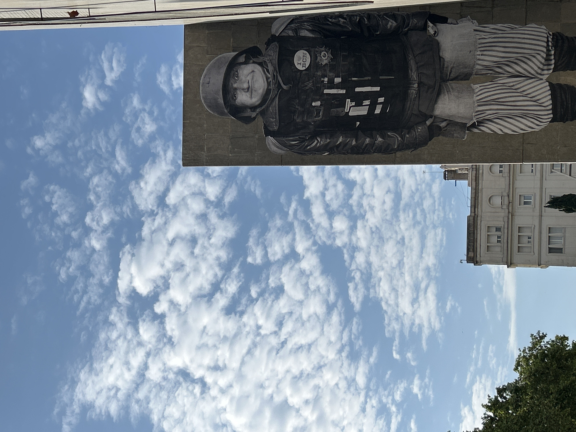 This outdoor image depicts a partially cloudy blue sky in Barcelona, Spain, positioned above a building. A mural of a person wearing a helmet, vest, and striped pants is on the building. The mural includes a button with the text "I heart BCN". In the bottom right corner of the image is a building with windows and a green tree.