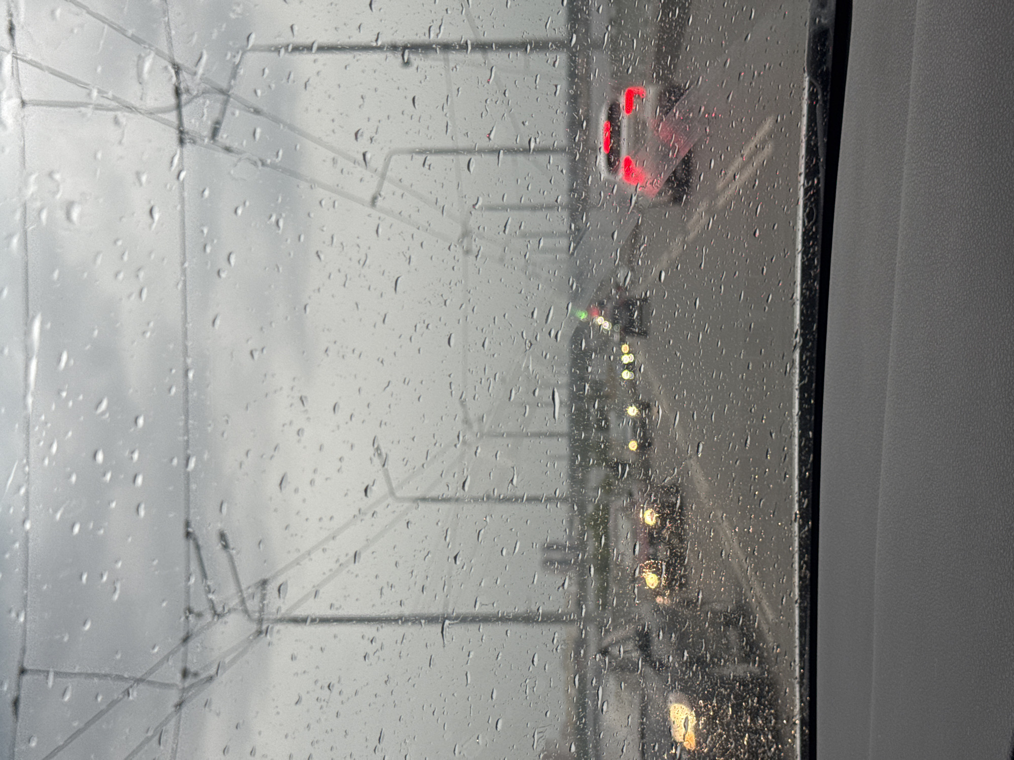 The image is a photograph taken through a wet window, likely a vehicle's windshield or a train window, showing a view of a road and overhead electrical wires. The window is covered in numerous raindrops, distorting the view and creating a diffused, atmospheric effect.
The scene depicts a highway or a similar roadway with several cars visible in the distance. One car in the middle ground has its red taillights illuminated. The road appears wet, consistent with the rain. Overhead, a network of electrical wires and poles is prominent, suggesting a tram line or an electric railway. The sky is overcast and grey, indicating a rainy or stormy day. The overall mood is somber and slightly melancholic, due to the weather and the blurred, desaturated colors.
The photograph appears to be taken during daylight, but the heavy cloud cover makes it appear dim. There are no people clearly visible, only the indistinct shapes of vehicles. The setting seems to be an urban or suburban environment, given the presence of electrical infrastructure and the roadway. No visible text is present in the image. The reflection on the window suggests the viewer is inside a vehicle, possibly moving, given the perspective and the blurry nature of the background. The texture of the interior of the vehicle is visible on the right side of the frame, appearing to be a dark, textured surface.