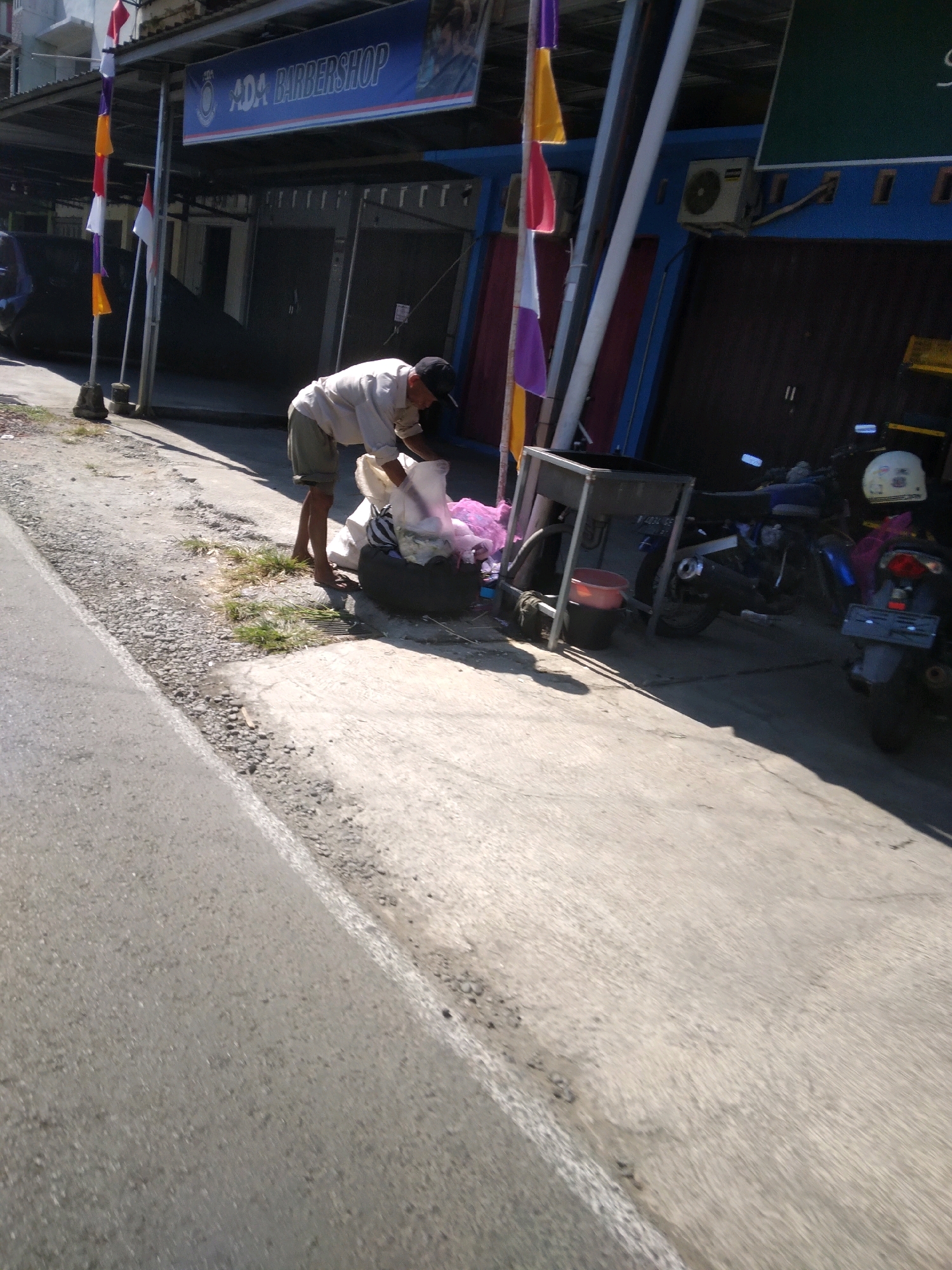 A man wearing a light-colored short-sleeved shirt and khaki shorts, and a black cap, is bent over a large, dark-colored garbage bag on a concrete sidewalk. He appears to be sorting through or gathering items from a clear plastic bag that is partially filled with white material. Next to the garbage bag is a metal stand or cart with a red bucket beneath it. In the background, a blue building has a sign overhead that reads "ADA BARBERSHOP." Several motorcycles are parked in front of the building, some with helmets visible. Flags in red, white, yellow, and purple are displayed on a pole to the left of the barbershop. The scene is outdoors, with a road visible in the foreground. The lighting suggests it is daytime. The location is identified as Karangsoka, Indonesia.