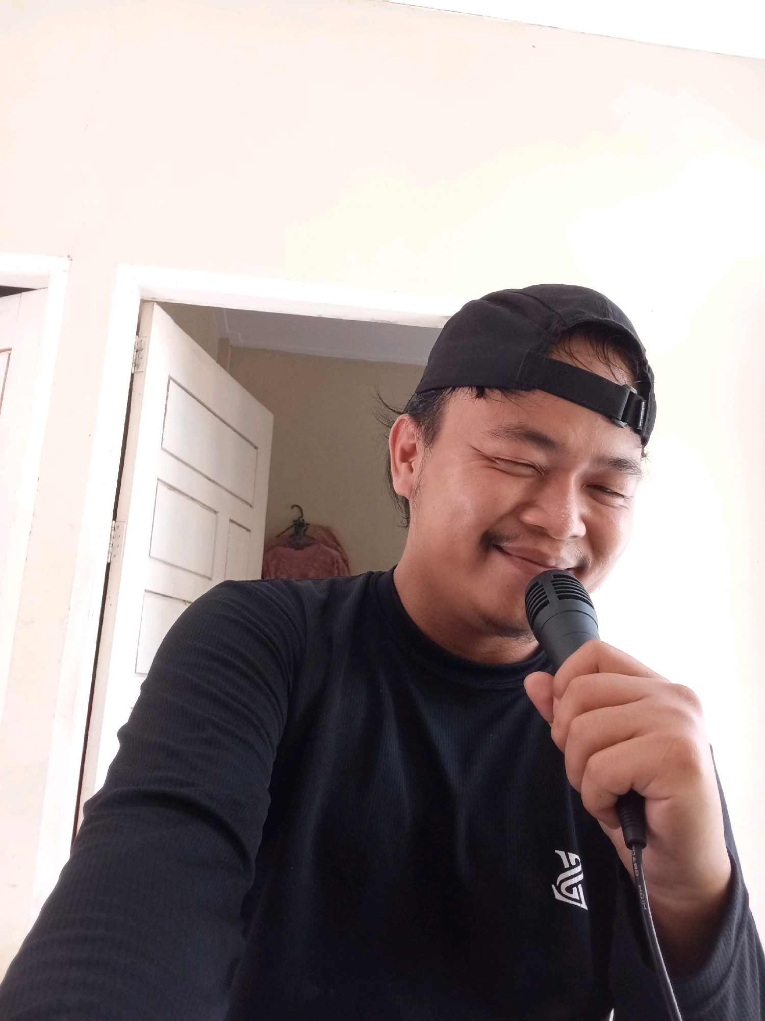 The image shows a young man wearing a black t-shirt and a black baseball cap, holding a black microphone to his face with his right hand. He has his eyes closed and a smile on his face. He is standing in front of a white door. The room appears to be a home interior, possibly in Kualu, Indonesia, judging by the style of the door and the lighting. The man seems to be singing or speaking into the microphone.