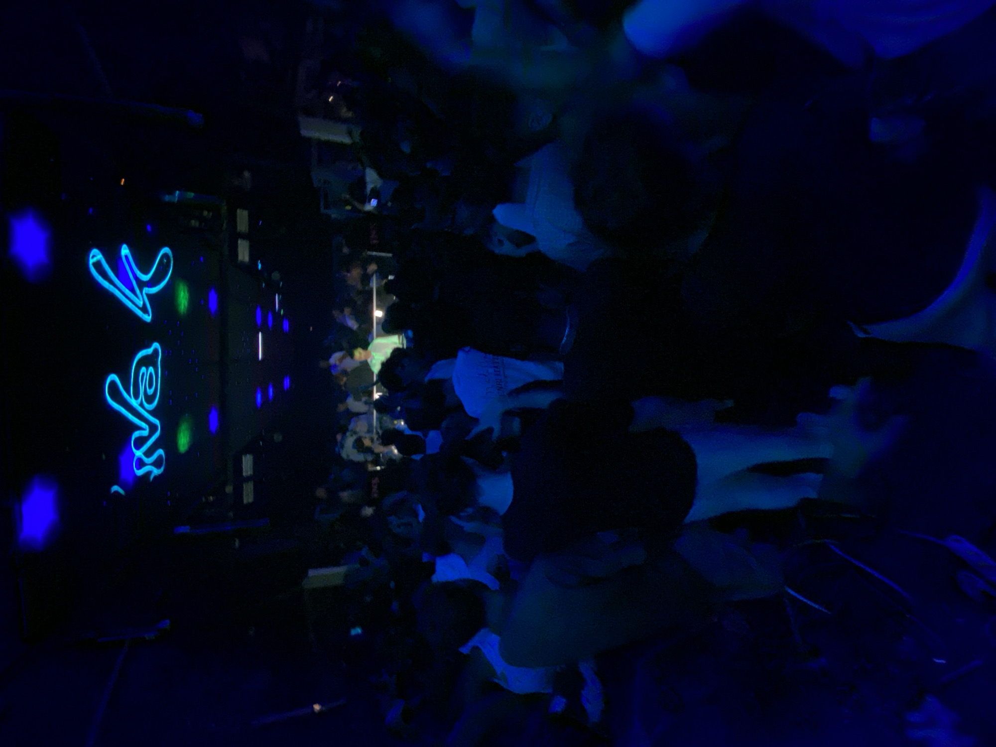 The image shows a crowded nightclub in Jesolo, Italy. A neon sign with blue lights that reads "Na k" is visible at the top of the frame. The nightclub is dimly lit, with blue and green lights illuminating the scene. A crowd of people is dancing and socializing in the space, with most of them dressed in casual attire. The overall atmosphere appears to be lively and energetic, characteristic of a typical nightclub scene. The perspective is from within the crowd, looking towards the stage or DJ booth area.