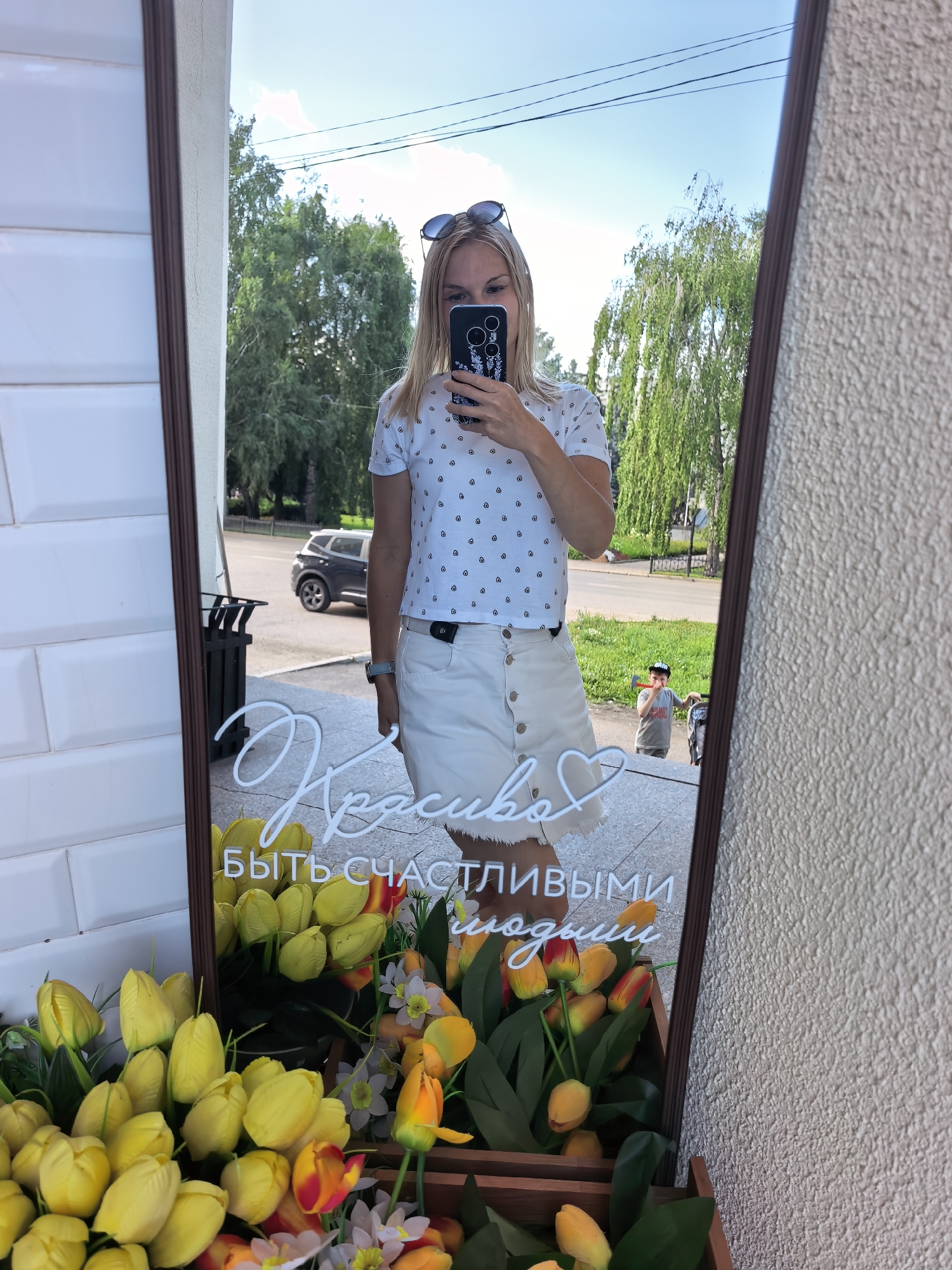 A woman with blonde hair and sunglasses is taking a selfie in a full-length mirror. She is wearing a white t-shirt with a black polka dot pattern and a white denim skirt. In the reflection, a street scene is visible with trees, a car, and a child holding a toy. The mirror is framed with dark wood and is placed in front of a wall with white tiled bricks on the left and a textured wall on the right. In the lower part of the mirror, there is a bouquet of yellow tulips and other flowers. A handwritten inscription in white neon letters is also visible in the mirror, reading "Красиво быть счастливыми людьми" which translates to "Beautiful to be happy people". The overall scene suggests an outdoor setting, possibly near a shop or cafe.