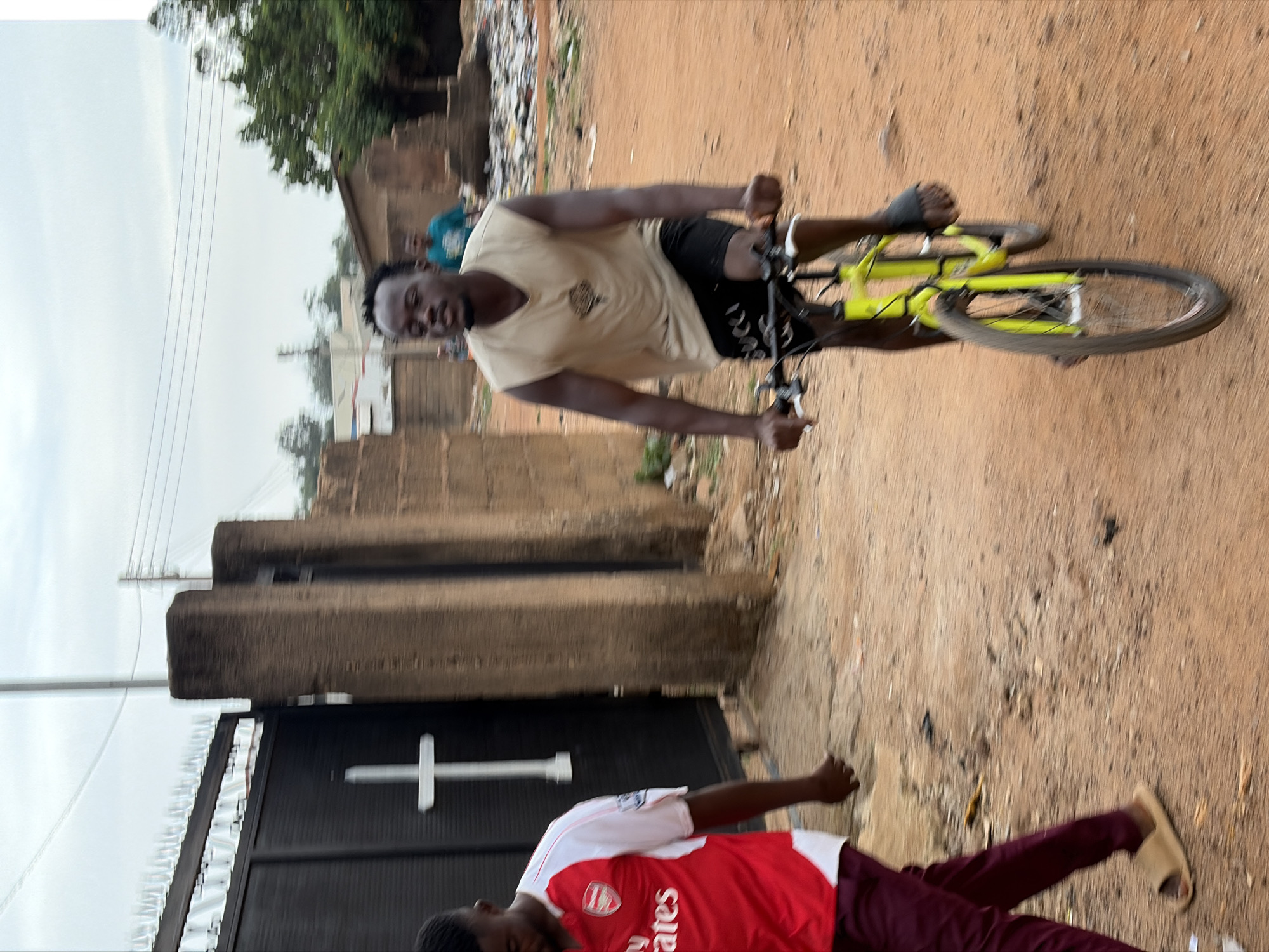 A man in a tan shirt and black shorts is riding a yellow bicycle down a dirt road. Another man wearing an Arsenal soccer jersey and maroon pants is walking in the opposite direction near a black gate with a white cross. The location appears to be Barakin Lerawi, Nigeria. The area is visibly poor and dusty, with debris and unpaved surfaces. Buildings are made of mud bricks.