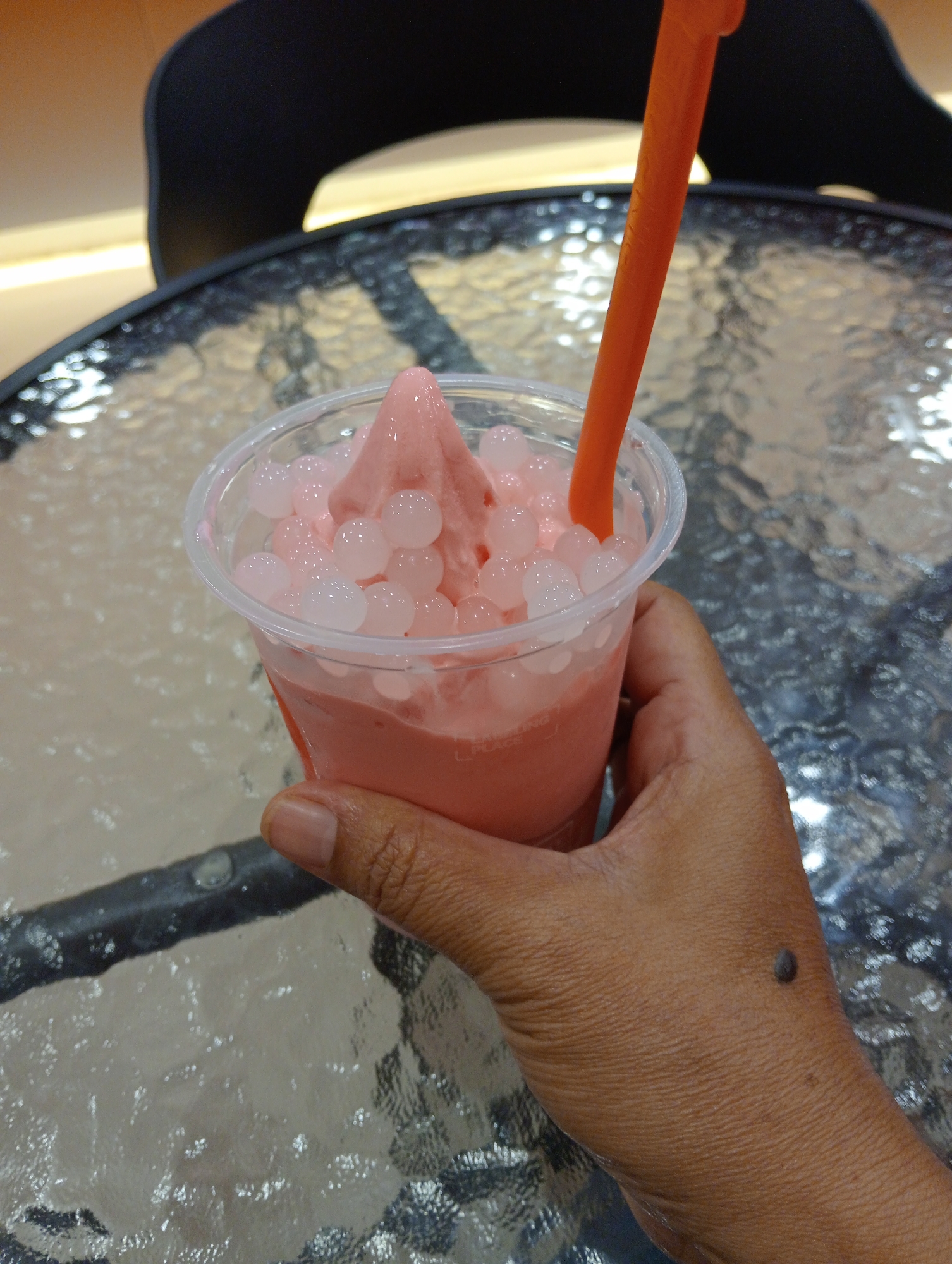 A close-up, eye-level shot shows a hand holding a clear plastic cup filled with a pink, blended drink topped with white boba pearls. An orange plastic straw protrudes from the drink. The cup is placed on a reflective, textured glass table, and a black chair is visible in the blurred background. The image appears to be taken in a café or similar establishment. The hand has a dark mole on the back. The overall scene suggests a casual refreshment. It is not possible to determine the specific location in Pringsewu, Indonesia, from the image alone.