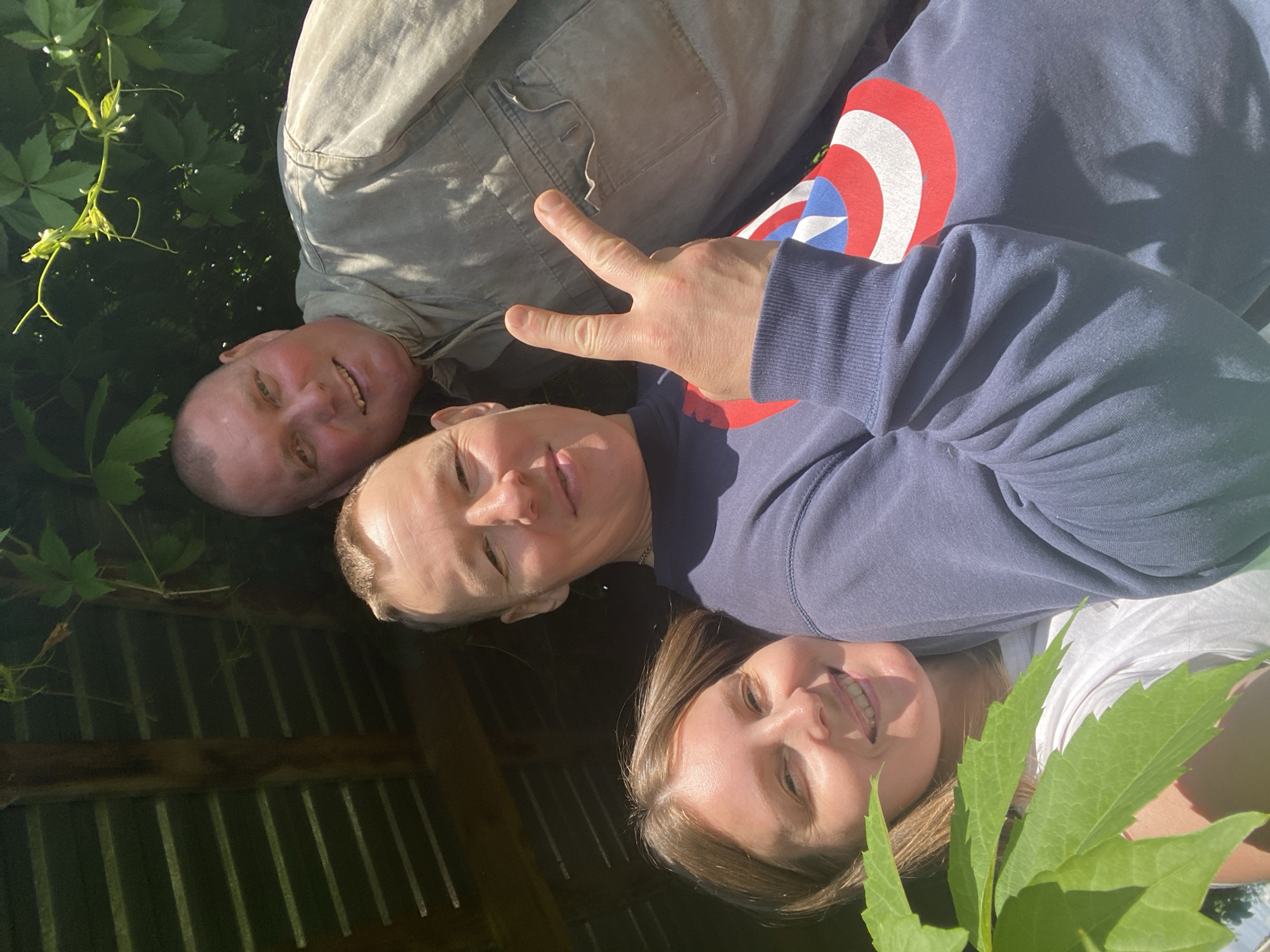 This group portrait features three individuals closely arranged, appearing to be lying down outdoors and looking up at the camera. The image is taken in bright daylight, casting visible shadows and illuminating their faces.
On the left, a man with short, light hair and a light-colored, possibly khaki, long-sleeved shirt is smiling broadly. His face shows a healthy, sun-kissed complexion. In the center, another man with short, light hair is giving a slight smile and making a 'peace' or 'V for victory' sign with his right hand. He is wearing a dark blue long-sleeved sweatshirt adorned with a prominent red, white, and blue Captain America shield logo on his left shoulder/chest. To the right, a woman with long, light brown hair is smiling widely, wearing a white t-shirt.
Green foliage, including leaves and vines, is visible on the left and bottom right edges of the frame, indicating a natural, outdoor setting. Behind the subjects' heads, a dark background with vertical ridges, possibly a corrugated metal roof or wall, and some wooden beams suggests they might be under a shaded outdoor structure.
The overall mood is joyful and relaxed, conveyed by the smiles on all three faces and the friendly gesture of the peace sign. The clear lighting suggests a sunny day. There is no other visible text apart from the Captain America logo. The scene depicts a casual, happy moment shared between friends or family outdoors.