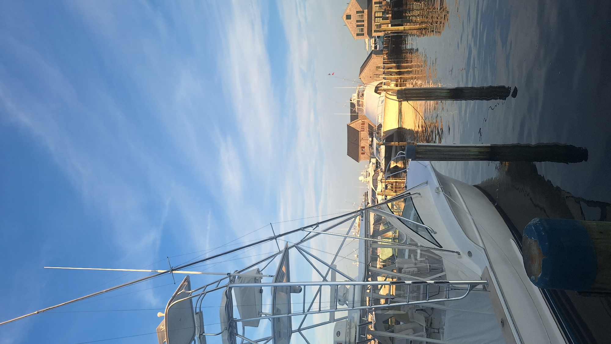 This is a vertical, outdoor, daylight photograph taken at the Nantucket Harbor in Nantucket, Massachusetts, United States of America. The image captures a marina scene with several boats docked and a clear blue sky with wispy white clouds.
In the foreground and left side of the image, a large white sport fishing boat is prominently featured. It has a prominent tower structure with multiple levels, antennas, and a canvas covering. The boat's hull is white with a blue stripe along the waterline. Part of its windshield and interior seating are visible. Adjacent to the boat, in the lower left, is a blue and weathered wooden post partially submerged in the water.
The water in the foreground is dark and reflective, showing the dark reflections of wooden pilings and the boat. Two wooden pilings stand vertically in the water, to the right of the foreground boat.
In the mid-ground, several other boats are docked. To the right, a white yacht with a sleek design is visible, reflecting the warm light of the setting sun. Behind it, wooden buildings with pitched roofs and weathered siding, characteristic of historic New England architecture, are visible along the waterfront. These buildings suggest a commercial or residential area adjacent to the harbor.
The sky is a bright blue with thin, wispy clouds streaking across it, indicating fair weather. The overall lighting suggests it is either early morning or late afternoon, with the sun casting a warm glow on the scene. The composition draws the viewer's eye from the large boat in the foreground towards the buildings and boats in the background. There are no people visible in the photograph.