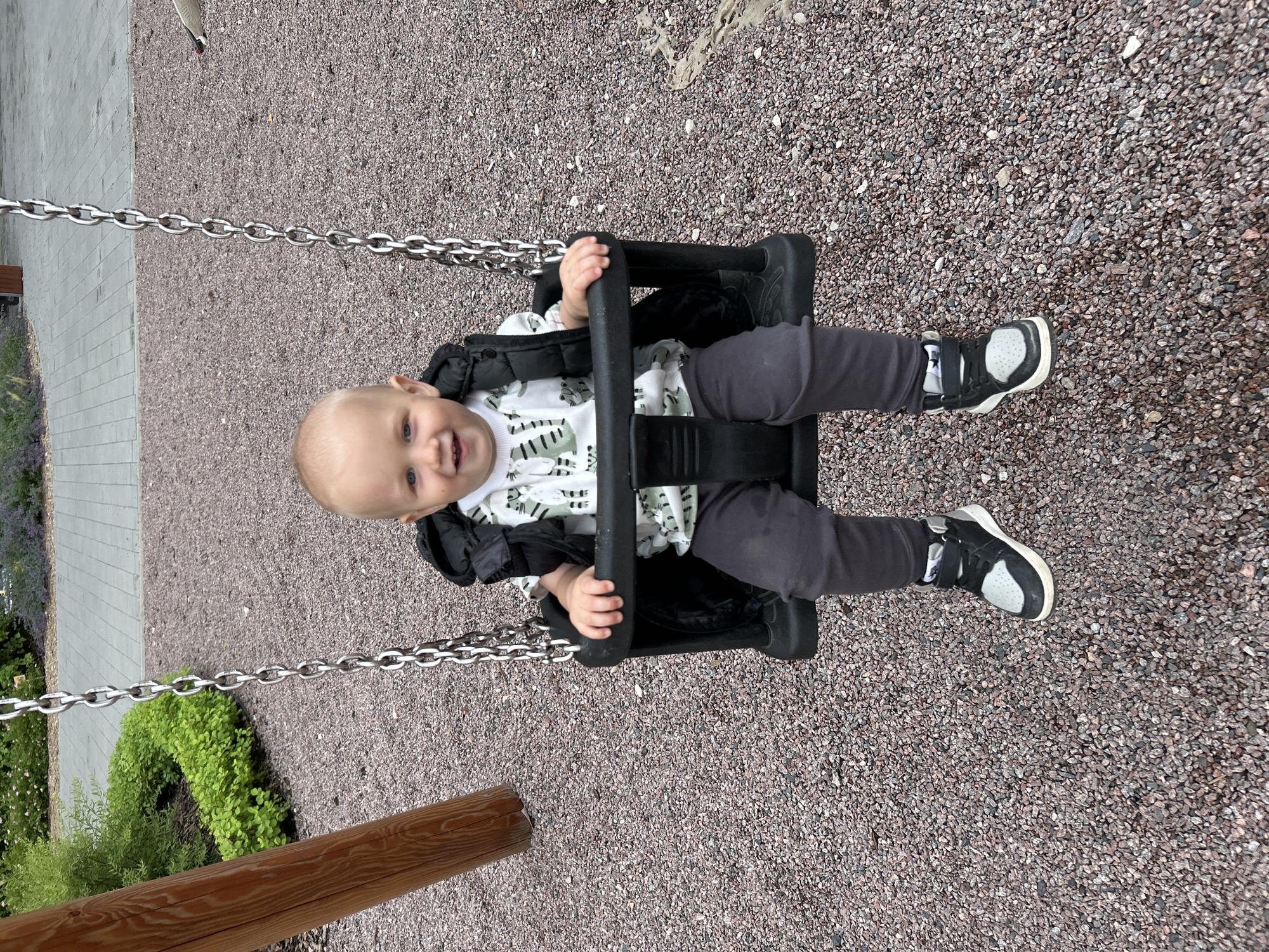 A young Caucasian child with blonde hair and blue eyes is sitting in a baby swing. The child is wearing a black vest over a white t-shirt with a green pattern and dark gray pants, with black and gray sneakers. The swing is black and attached to silver chains. The child is smiling. The background consists of a gravel-covered playground with a wooden support post on the left and some greenery and paved walkways in the distance. The location is a playground in Kaluga, Russia.