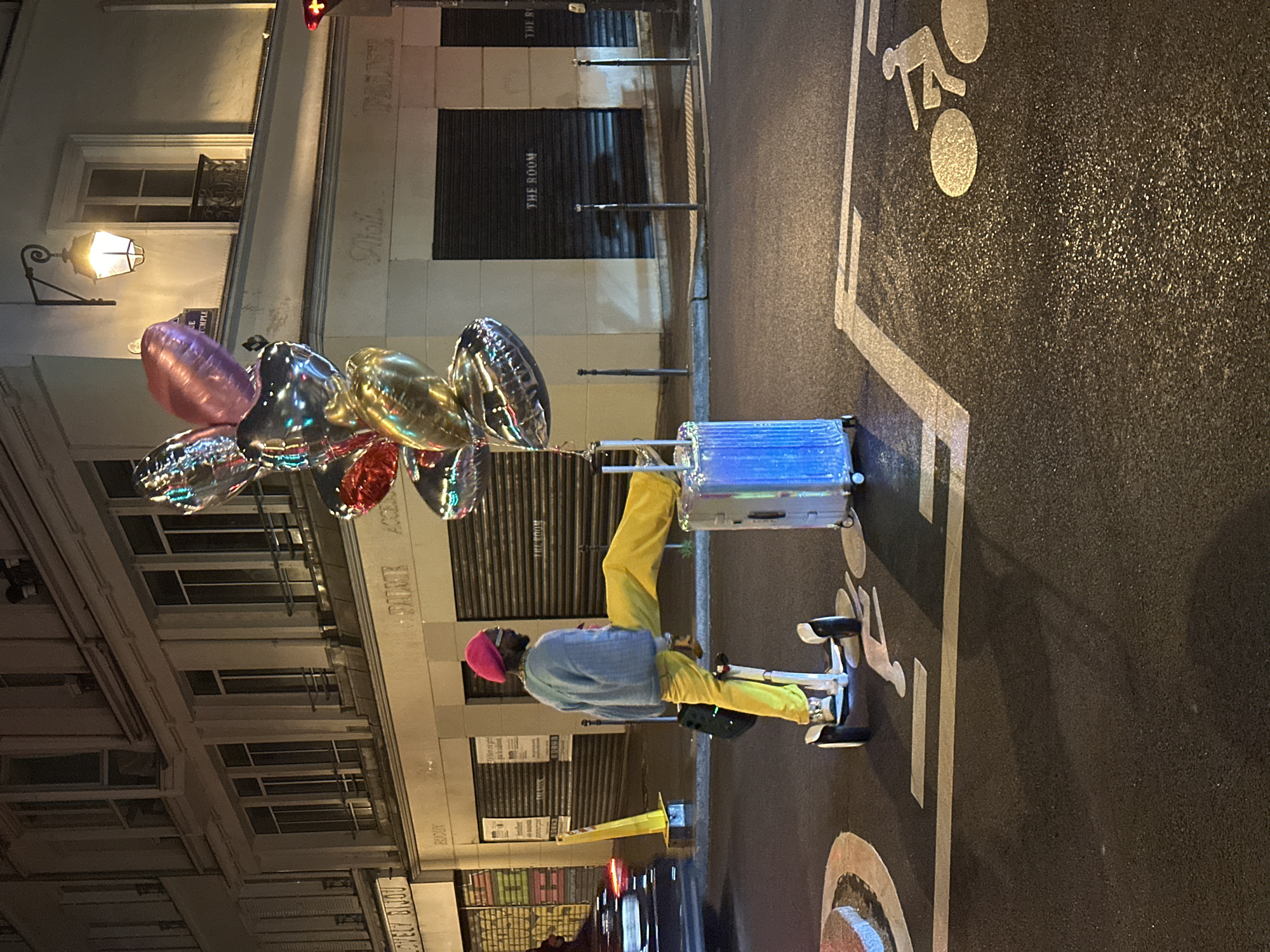 An individual is depicted at night on Rue du Temple in Paris, France, balancing on a white two-wheeled self-balancing scooter (hoverboard-type device). The person, appearing male, wears a bright pink beanie, a light blue textured sweater, and bright yellow loose-fitting pants, with a dark beard. Their left leg is extended forward, resting on a prominent, upright bright blue corrugated hard-shell suitcase with an extended handle. The suitcase supports a large cluster of approximately 8-10 metallic, heart-shaped balloons in various colors including silver, gold, pink, and red, reflecting ambient light. The individual carries a black bag over their left shoulder.
The scene unfolds on a wet asphalt street, illuminated by artificial light sources. Road markings, including a white solid line, a white bicycle symbol, and a white stop line, are visible on the street surface. In the background, typical Parisian architecture features multi-story buildings with windows and decorative elements. Storefronts at street level are closed with security shutters, some displaying "THE ROOM" and "LOVELY BIJOU." A yellow traffic cone is present in the left background, and a street lamp projects light from a building facade on the right. A faint light reflection from a vehicle is visible in the far left.