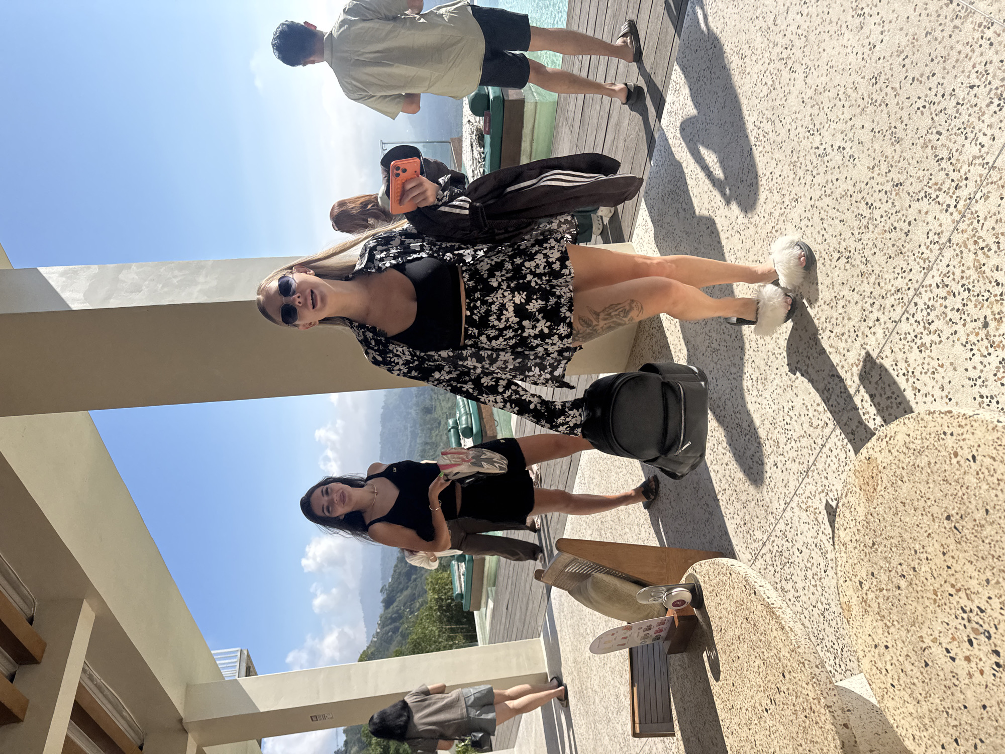 In Kintamani, Indonesia, a group of people are gathered on a wooden deck near a swimming pool. A woman in the foreground, wearing a black crop top, a floral-print skirt, a matching open shirt, and fluffy white slippers, is holding an orange smartphone in her right hand. She is also carrying a black jacket with white stripes over her left arm and a black backpack at her feet. To her left, another woman in a black dress and sandals is holding a patterned bag. In the background, a man in a light-colored shirt and dark shorts stands near the pool, and a woman with red hair is visible between the two women in the foreground. The scene is set against a backdrop of lush green hills under a clear blue sky.