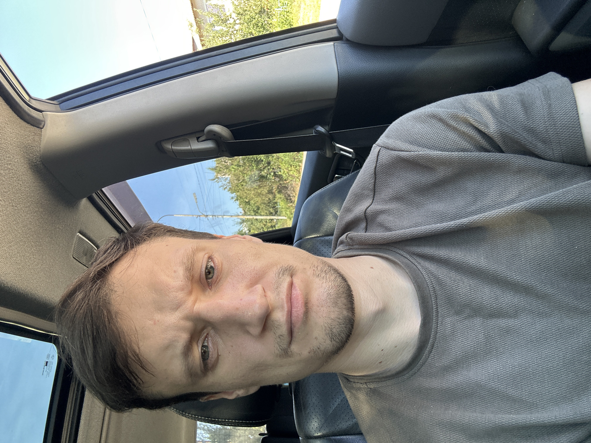 A man is captured in a selfie from the chest up, seated inside a vehicle. He has brown hair and is wearing a grey t-shirt. His expression appears to be one of concern or skepticism, with his eyebrows furrowed and his mouth slightly downturned. The interior of the car is visible, including a black leather seat and a portion of the dashboard. Sunlight streams in through the open window and sunroof, indicating it is daytime. The background shows greenery and a building, suggesting an outdoor setting in a suburban or rural area. The location is identified as Makiv, Ukraine.