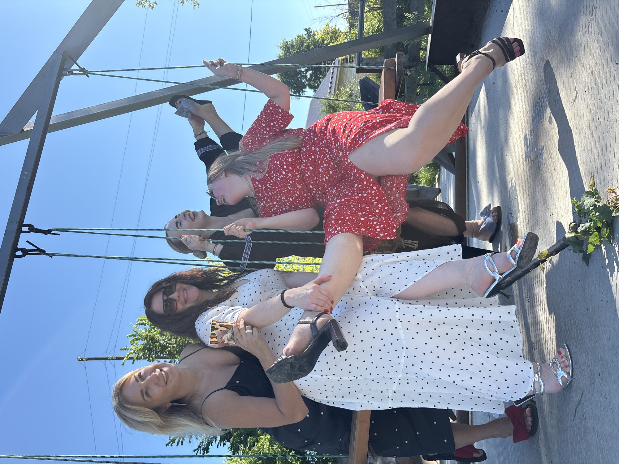 Four women are posing for a photograph on a swing set on a sunny day. The woman on the far left is wearing a black polka dot dress and black heels. She is holding a glass of champagne. The woman next to her is wearing a white polka dot dress and silver heels. The third woman from the left is wearing a black patterned dress and is laughing. The woman on the far right is wearing a red polka dot dress and is also laughing. They are all on a swing set, with two women on the swing and two standing behind them. The background shows a clear blue sky and some trees. The scene appears to be in an outdoor area, possibly a backyard or park. The city of Zav'yalovo, Russia, is mentioned as the location, which suggests a rural or suburban setting.
