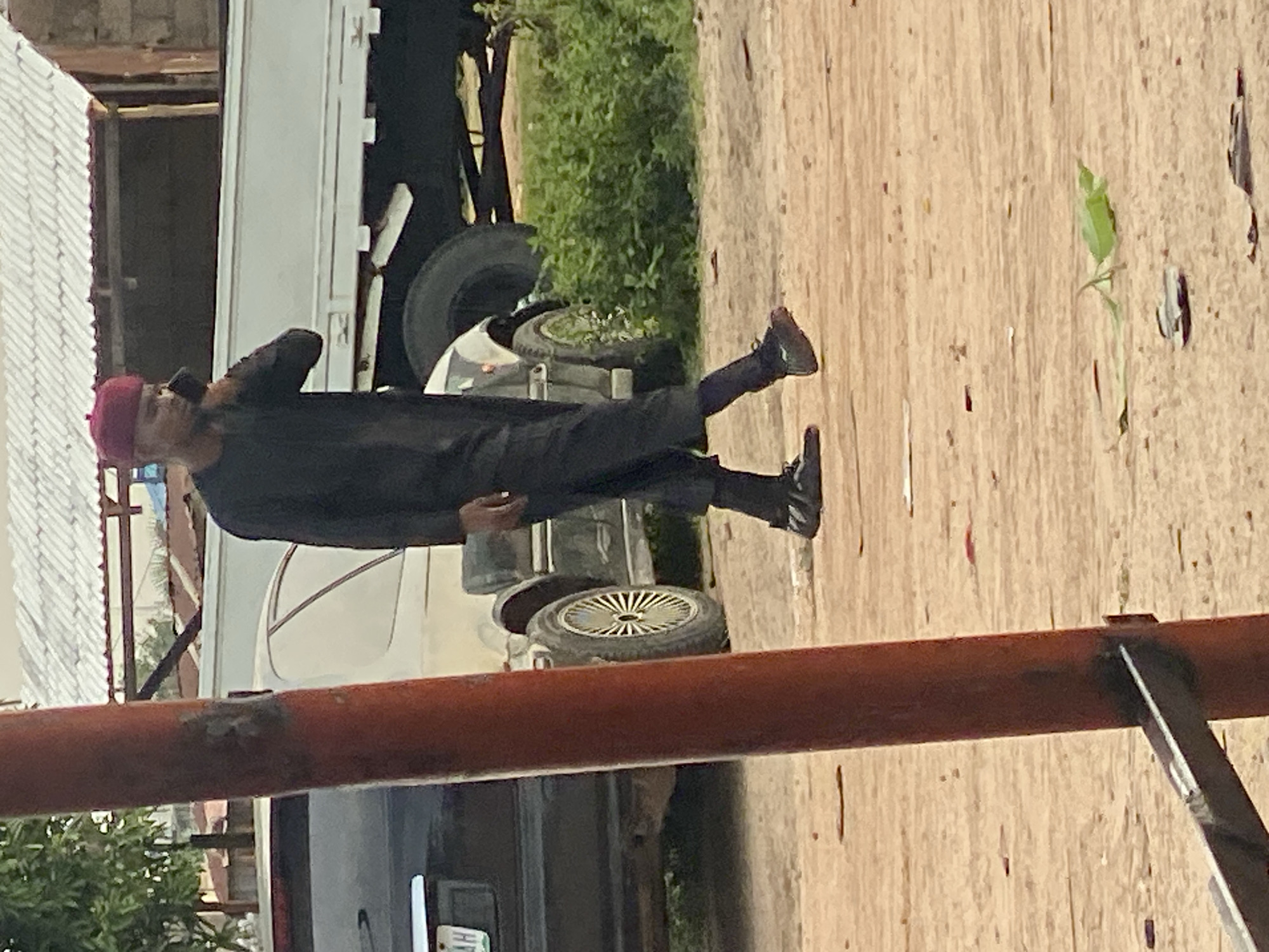 A young man wearing a black robe and a maroon cap is walking on a dirt road in Jalingo, Nigeria, while talking on a cell phone. He is positioned in the mid-right of the frame. In the background, a white truck is parked, and to the left of that, a dark sedan is partially visible. A large, orange-red metal pole is in the foreground, obscuring the left side of the image. The environment appears to be an outdoor, possibly semi-urban or rural area, with some vegetation visible behind the truck. The road surface is dusty and uneven.