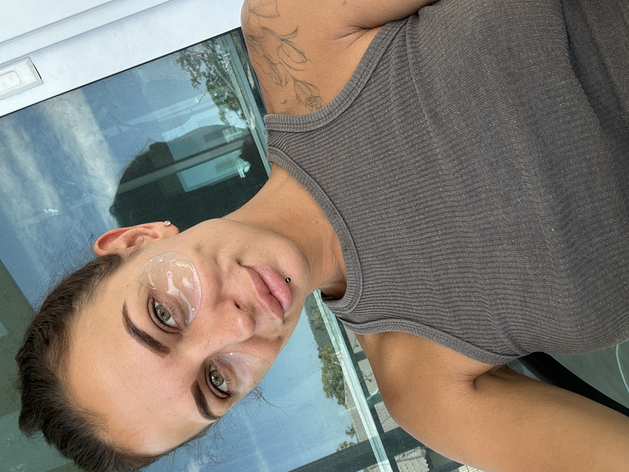 A woman is taking a selfie with an eye mask on, while sitting near a window overlooking a tropical landscape. The woman has dark hair pulled back in a bun, green eyes, and a nose ring. She is wearing a grey tank top. Her left arm has a floral tattoo. The window appears to be in a room in Ko Samui District, Thailand, as suggested by the tropical trees and foliage visible through the glass. The reflection in the window shows the sky with clouds and more foliage.