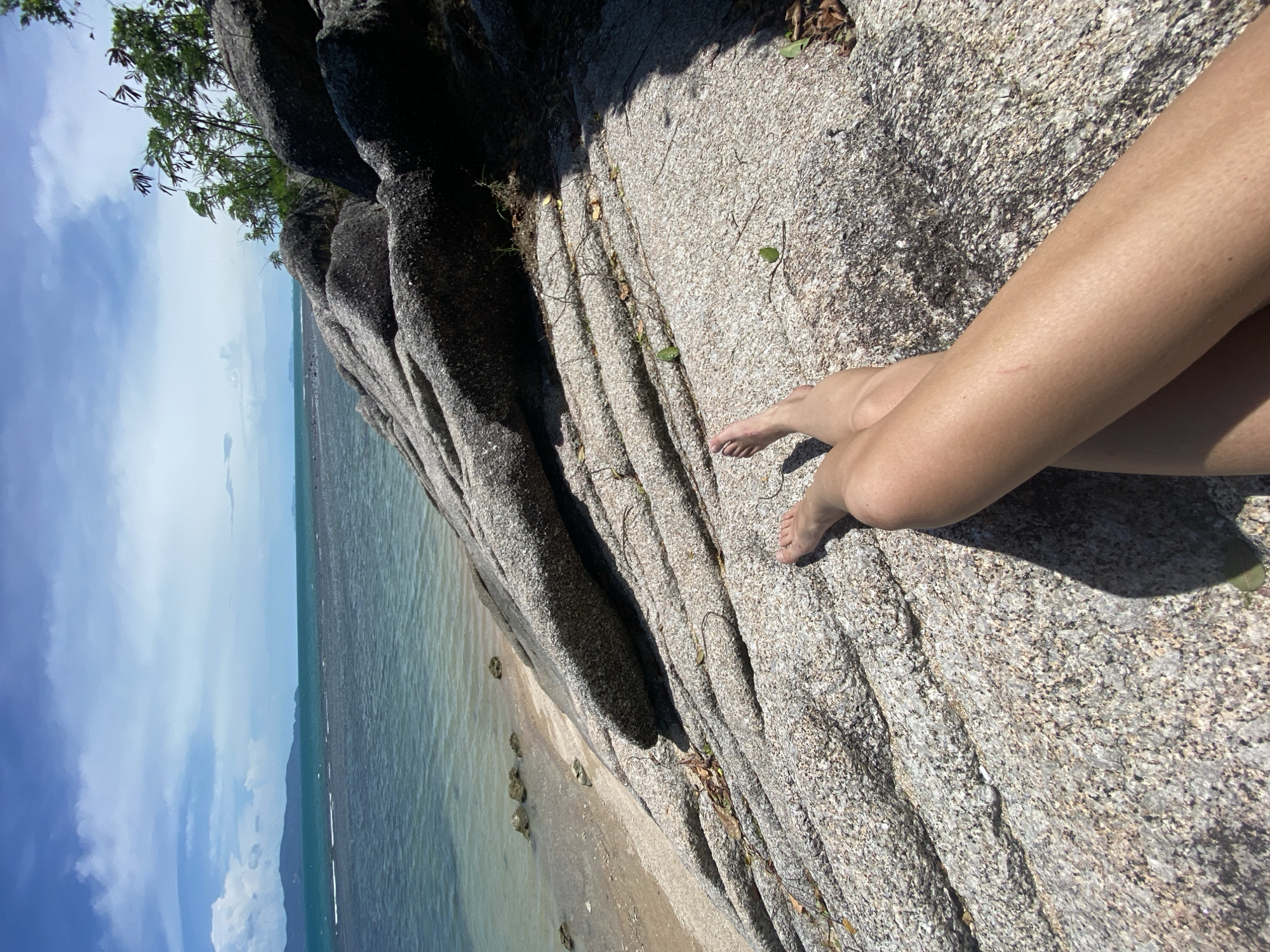 The image shows a person's legs crossed, resting on a large rock formation. The location is likely a beach, as evidenced by the turquoise water and sandy shoreline visible in the background. Dark gray rocks rise above the beach. Some trees are seen at the top edge of the scene. The sky is blue with some clouds. Based on the prompt location, this scene might be near Ban Haad Rin, Thailand. No other people are visible.