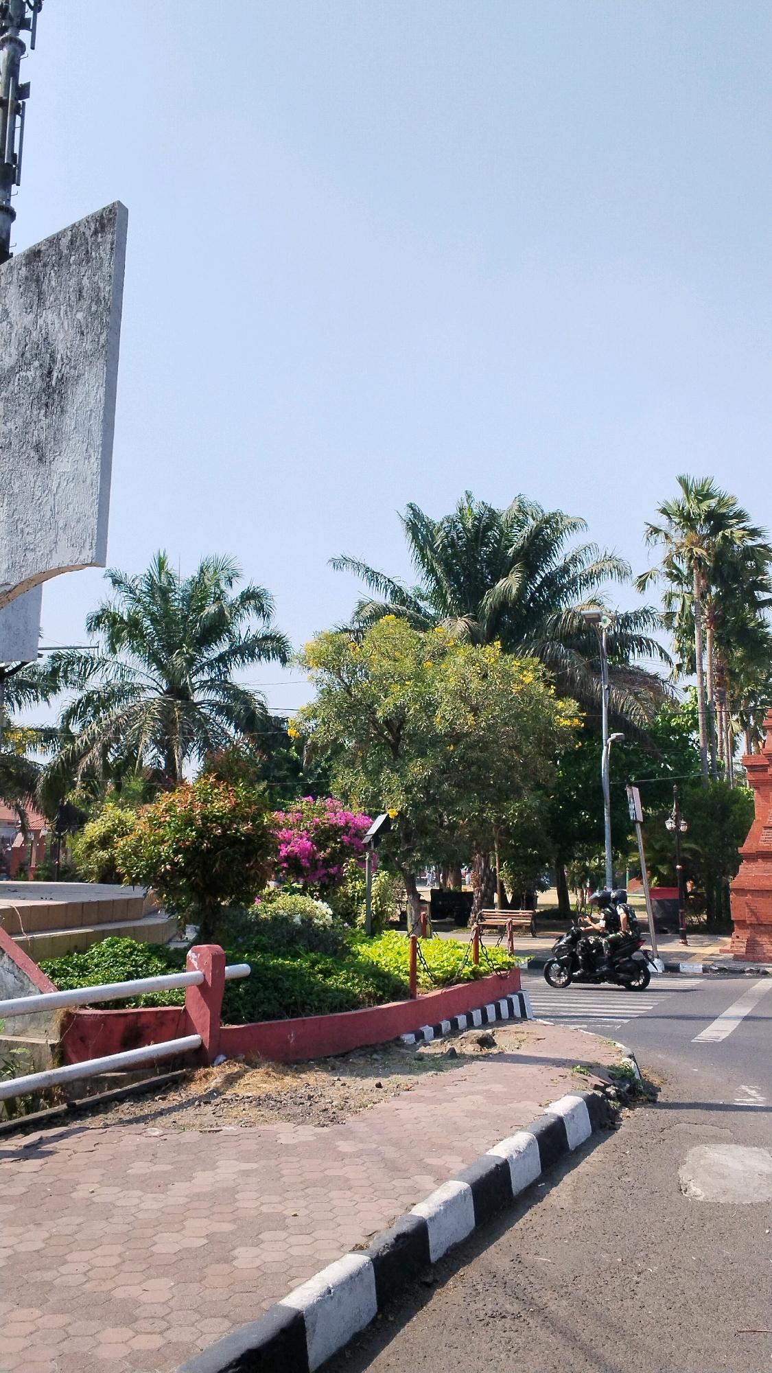 This image captures a street scene in the city of Mojokerto, Indonesia. On the left, a large, weathered concrete structure, possibly part of a monument or building, is partially visible. In the center, a well-maintained garden area features lush green bushes, flowering plants in vibrant pink, and a tree with yellow blossoms. Behind the garden, several palm trees and other trees create a dense canopy. A streetlight stands near the garden. A motorcycle with two occupants, wearing helmets, is traveling on the road in the background, crossing a pedestrian walkway. The walkway is paved with hexagonal bricks and bordered by a curb painted with alternating black and white sections. The overall environment appears to be a public space, likely a park or roadside area, under a clear blue sky.