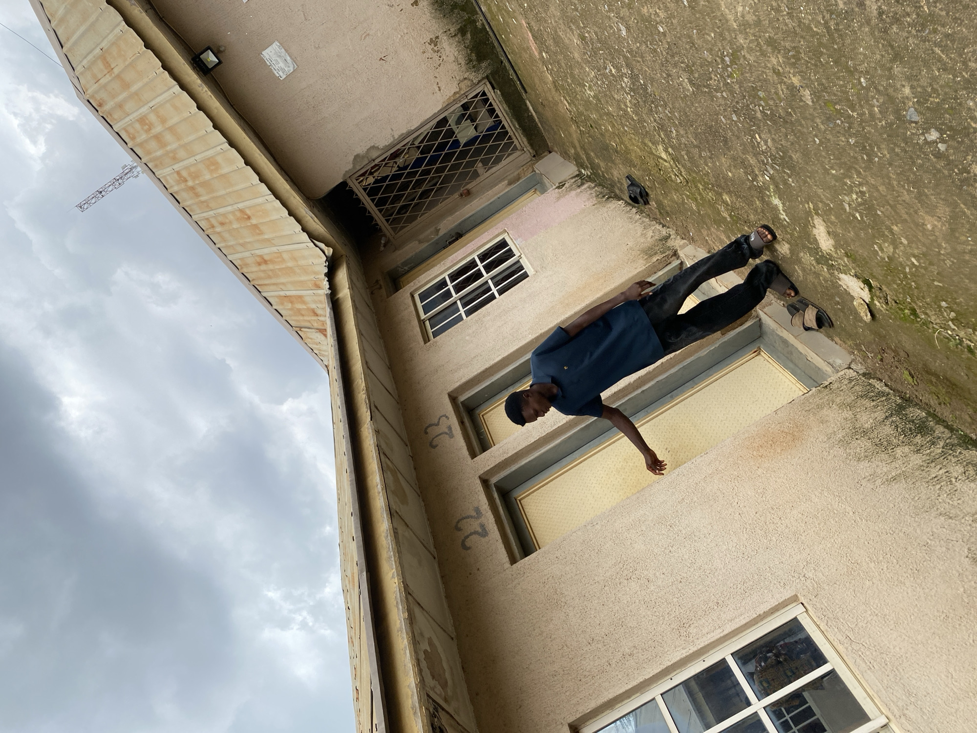 A young Black man wearing a navy blue polo shirt and dark pants stands on a concrete courtyard in front of a building in Jos, Nigeria. The building appears to be an apartment complex or a similar multi-unit dwelling. Two doors are visible, labeled with the numbers "22" and "23." The man is reaching out his right hand towards door number 22. The building has a corrugated metal roof, and the sky is overcast with dark clouds. A cell tower is visible in the distance. There are several pairs of sandals scattered on the ground around the building.
