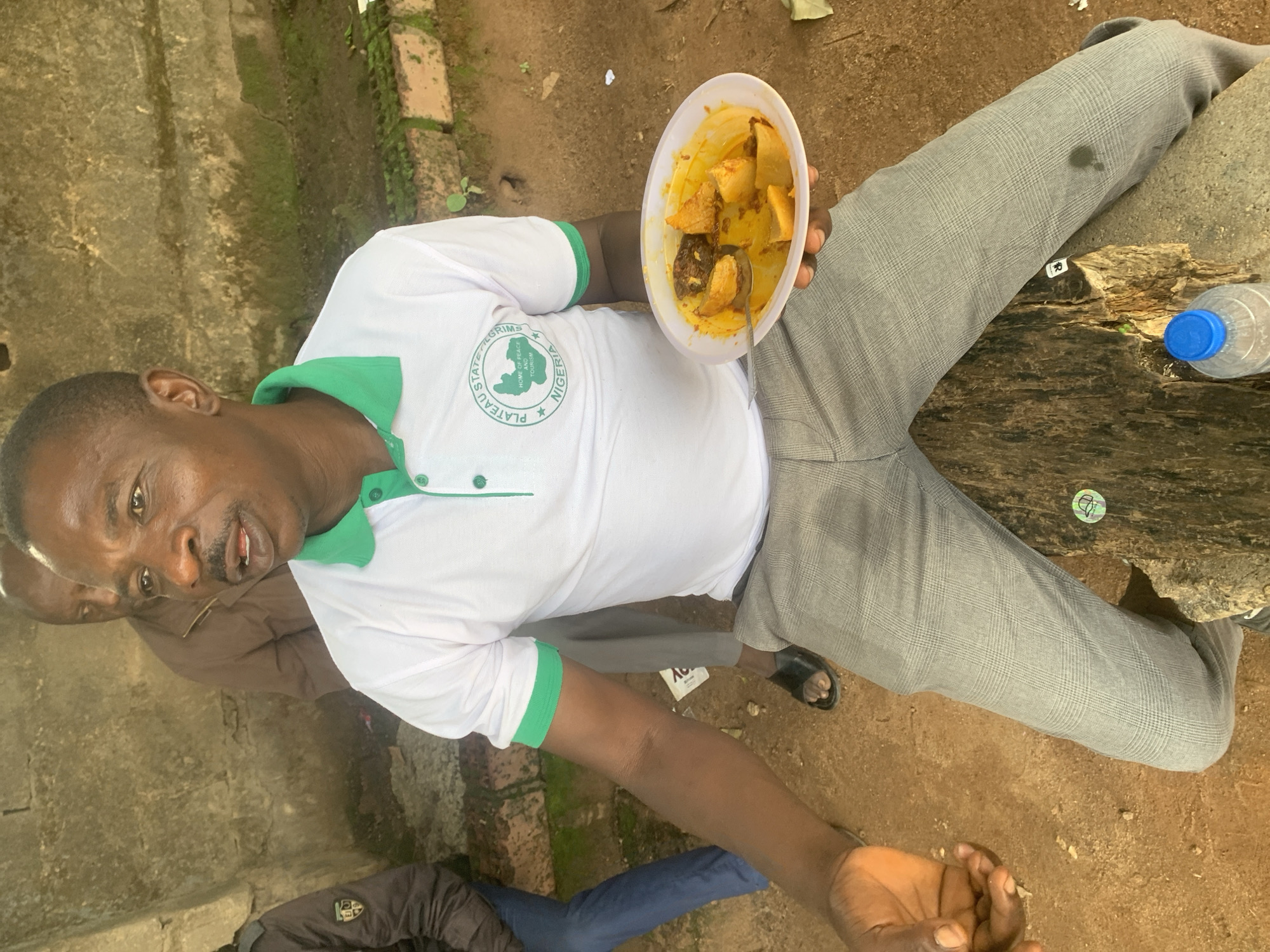 The image shows a man sitting outdoors, holding a white bowl of food with a spoon. He is wearing a white polo shirt with a green collar and trim, and grey checkered trousers. The shirt has a logo that appears to be circular with a map outline and text. There is a water bottle with a blue cap to his right, placed on a rough wooden surface. The setting appears to be a rustic outdoor area, possibly with some brickwork and dirt ground. Another person is partially visible behind him, and the lower half of another person's legs wearing blue pants are visible in the foreground. The lighting suggests it is daytime, and the ground appears dry, possibly indicating a dry weather condition. The man's expression is neutral. The logo on his shirt has text that seems to include "PLATEAU STATE", "HOME OF PEACE AND TOURISM", and "NIGERIA". A sticker is visible on the wooden surface, also appearing to be circular with a symbol.