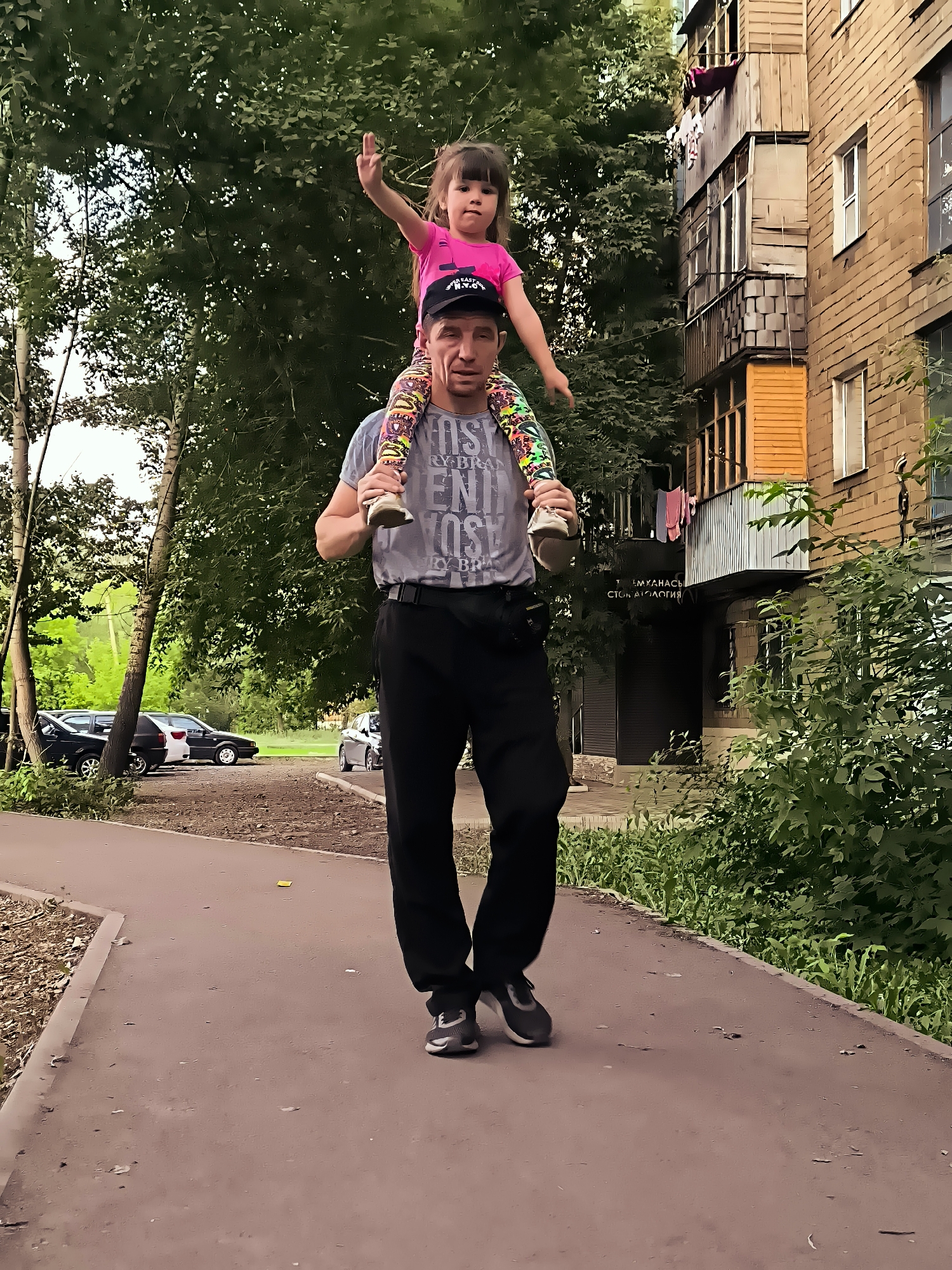 A man walks forward carrying a young girl on his shoulders. The man is wearing a grey t-shirt with text, black pants, and black and grey athletic shoes. The girl is wearing a bright pink t-shirt and colorful leggings. She has her right arm raised. They are walking on a paved path. To the left of the path, there is a dirt area with some parked cars and trees. To the right of the path is a brick apartment building with balconies and windows. The environment appears to be urban, with trees and vegetation present.