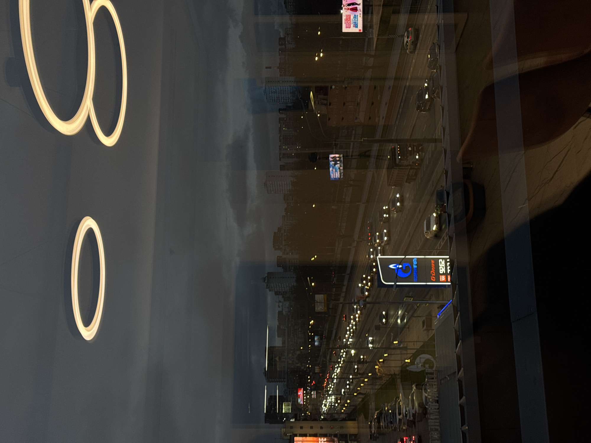 The image depicts a view from a high vantage point, likely an apartment or office building, overlooking a cityscape in Moscow, Russia. The view is seen through a glass window, causing reflections of interior elements such as light fixtures, possibly decorative circular lighting elements attached to the wall and indistinct human figure.
Outside, a major thoroughfare or highway is visible with numerous vehicles moving along it. Streetlights illuminate the roadway, creating lines of light stretching into the distance. The reflection of buildings, including high-rise structures, can be seen in the window, superimposed over the city view. A gas station with a large sign "G-Drive" is clearly identifiable. In the background, the sky is overcast. The image is taken during twilight or at night.