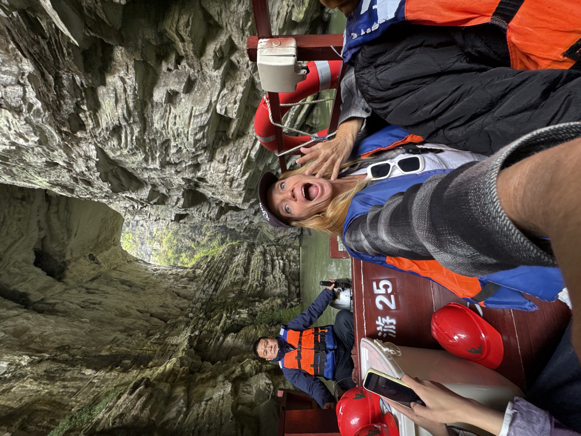 The image captures a boat tour passing through a narrow gorge in Zhuoshui, China. Passengers are wearing life jackets and red safety helmets. A woman in the boat, situated near the front, reacts with a surprised expression, while another passenger holds a phone, appearing to take pictures. The boat driver is at the helm. The gorge walls are composed of rock formations with openings visible at the end of the gorge. The boat's number, "25," is printed in both English and Chinese.