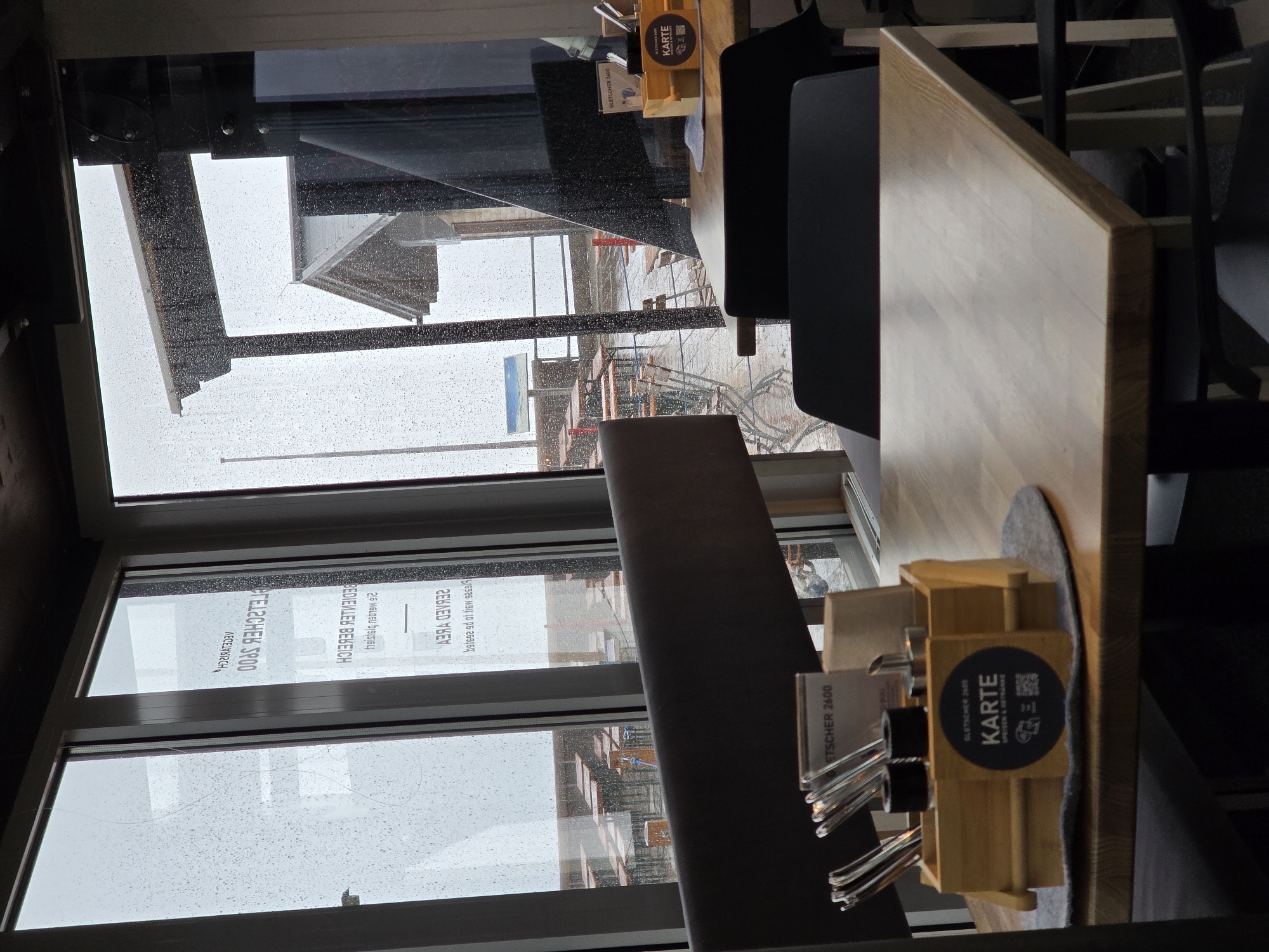 The image shows a view through a window with raindrops on it, looking out at an outdoor seating area with many tables and chairs. The tables are set and some have umbrellas. The outdoor area appears to be wet, suggesting it is raining or has recently rained. In the background, there is a building with a sloped roof. Inside the building, there are tables and chairs. On the table in the foreground, there is a wooden holder with cutlery and a sign that says "Gletscherr 2600 KARTE SPEISEN & GETRANKE." This indicates the location is likely a restaurant or cafe at an elevation of 2600 meters, possibly associated with a glacier. Garmisch-Partenkirchen is a town in Bavaria, Germany, known for its mountain resorts, so a restaurant at this altitude is plausible. The overall impression is of an indoor dining area with a view of an outdoor space during inclement weather.