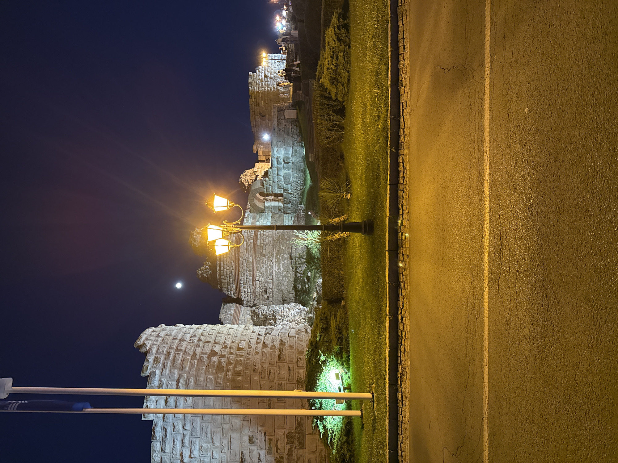 This is a nighttime, eye-level shot of an illuminated ancient stone fortress or wall in Nesebar, Bulgaria. The foreground shows a dark asphalt road with a white line, a cobblestone curb, and a strip of well-maintained green grass with bushes. A triple-headed lamppost, lit with warm yellow light, stands prominently in the middle ground. Behind the lamppost, the ancient stone structure rises, with sections of it illuminated by green lights at its base. Two flagpoles with flags are visible to the left of the structure. In the background, more of the ancient ruins are visible, with small groups of people walking around. The night sky is dark blue, with a bright full moon visible. The overall lighting creates a dramatic and historical ambiance.