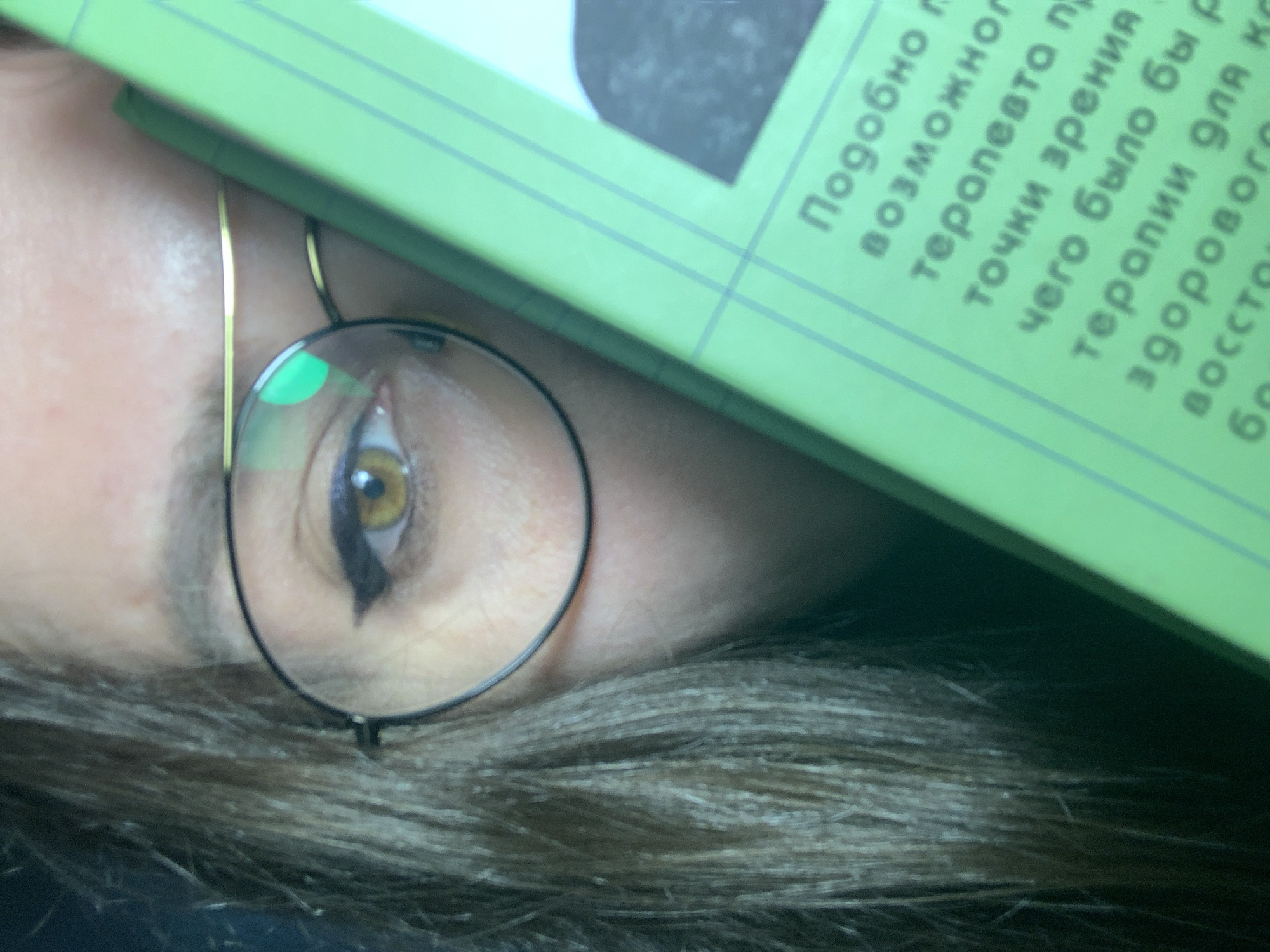 The image shows a close-up of a person's face, partially obscured by a green book. The person is wearing round glasses with thin black frames. The visible eye is hazel-colored and has dark eyeliner. Some hair is visible, appearing to be brown or grey. The book is positioned over the person's forehead and upper face, displaying some Russian text. The setting is Unknown, Unknown.