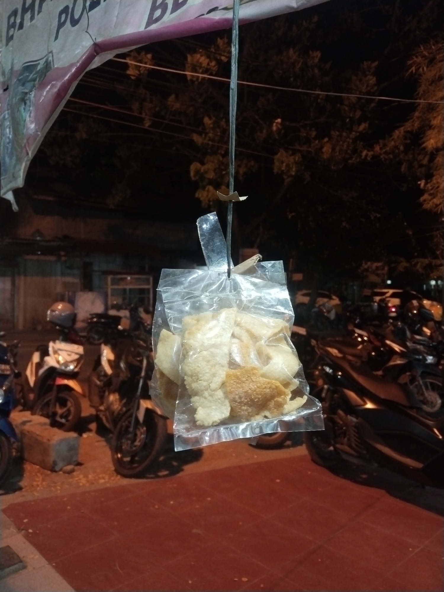 A bag of crackers hangs suspended in the air at night in Lhokseumawe, Indonesia. The crackers are visible within a clear plastic bag. The bag is hung from a thin metal rod. The surrounding area is tiled in red. Several motorbikes are parked in the background, along with other buildings. A banner with text is partially visible at the top of the frame. Dark trees are visible in the distance.