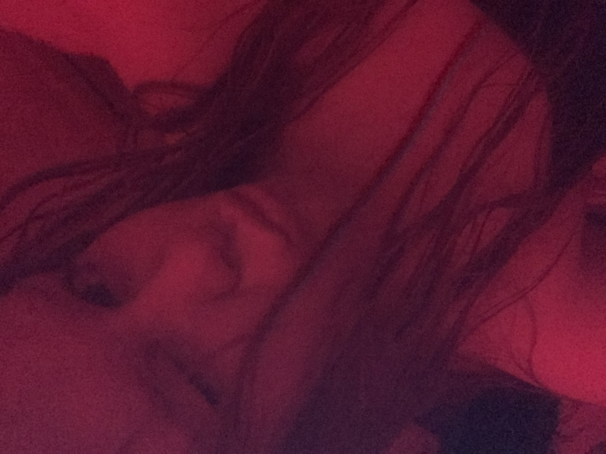 The image presents a close-up, dark, and blurry scene heavily filtered with a dominant deep red and maroon hue. A person, likely female, is partially visible, with long, dark hair cascading across the frame, obscuring much of their face and body. A faint outline of a cheek and possibly a portion of the nose or mouth can be discerned on the left side of the frame, emerging from the shadows.
The setting appears to be indoors, suggested by the artificial red lighting, which creates an intimate or subdued atmosphere. The background seems to consist of soft, red fabric, possibly bedding or an item of clothing, further indicating a private or relaxed environment, likely at evening or night. The person appears to be resting or lying down. Due to the low light, blurriness, and obscured features, specific emotions are not discernible. No distinct objects or visible text are present in the image. The exact location within Russia cannot be identified from the visual information provided.