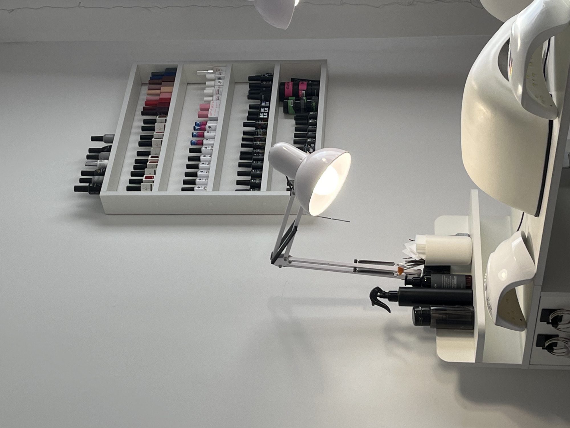 The image shows a section of a white wall. On the wall is a white shelf containing bottles of nail polish, arranged in color order. A white desk lamp is clipped to the shelf below, and its light is shining. To the right of the shelf is a larger white structure, with shelves holding various bottles and tools, and a nail lamp. The wall is featureless. There is no indication that the image was taken in Фоки, Russia.