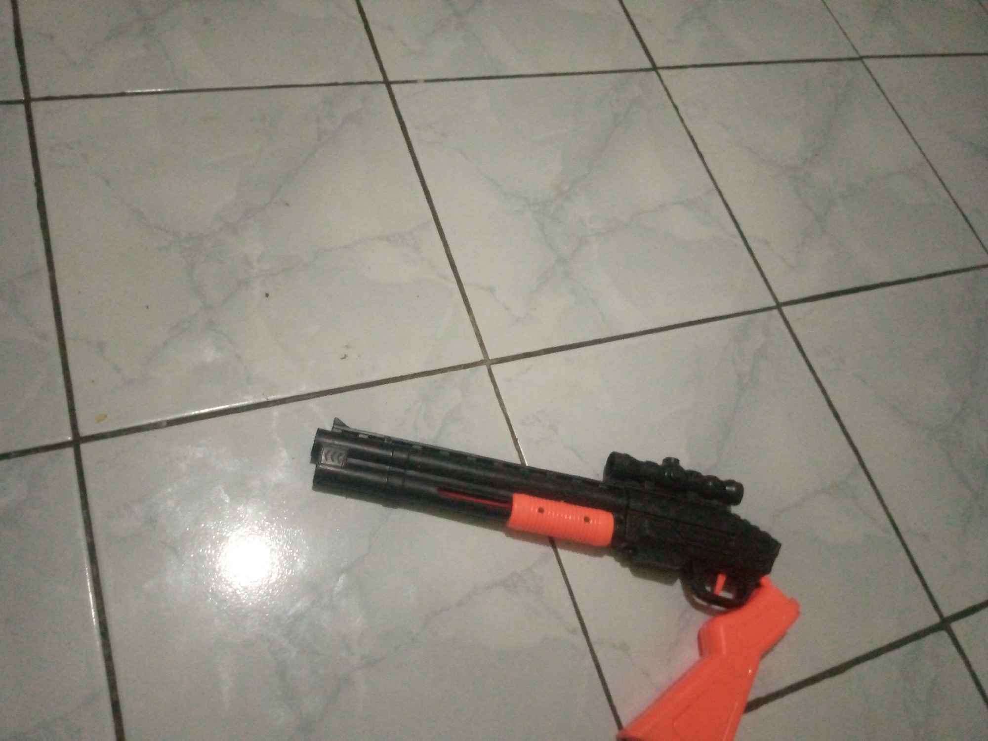 A toy shotgun with an orange stock and foregrip, and a black body with a scope, lies on a tiled floor. The tiles are light gray with darker gray grout lines forming a grid pattern. The lighting in the scene is somewhat dim, with a bright reflection on the floor near the toy. There are no people present in the image. The image appears to be taken indoors, likely in a residential setting. The location of Laksanamekar, Indonesia, is irrelevant to the visual content of the image.