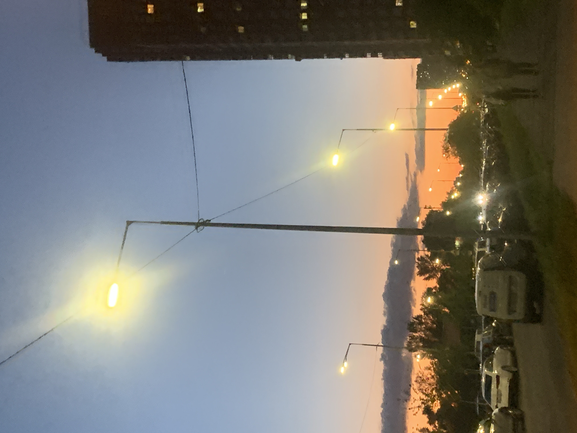 The image depicts a street scene at dusk in Kyiv, Ukraine, characterized by a gradient sky transitioning from a clear blue at the top to warm orange and pink hues near the horizon. Several streetlights are illuminated, casting a bright yellow glow. A tall, dark building, likely an apartment complex, is visible on the right side of the frame with its windows lit. On the left, a line of parked cars is present, including a white sedan and a white van. In the foreground, a paved walkway is visible, with two indistinct figures walking away from the viewer towards the right. Trees and foliage are present along the left side of the street, with some appearing silhouetted against the colorful sky. Power lines are strung across the scene, connecting to the lampposts. The overall impression is a typical urban evening in an apartment block district.