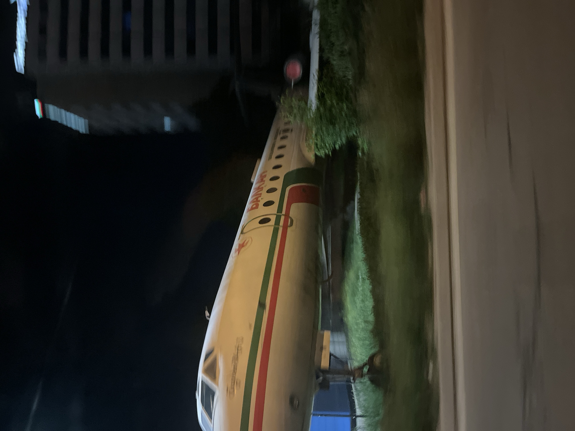 The image shows a grounded airplane fuselage with "Balkan" written on its side. The fuselage is white with green and red stripes. The background includes a dark night sky, a partially visible modern building, and greenery along the ground. It is at the Children's Science Center Muzeiko in Sofia, Bulgaria.