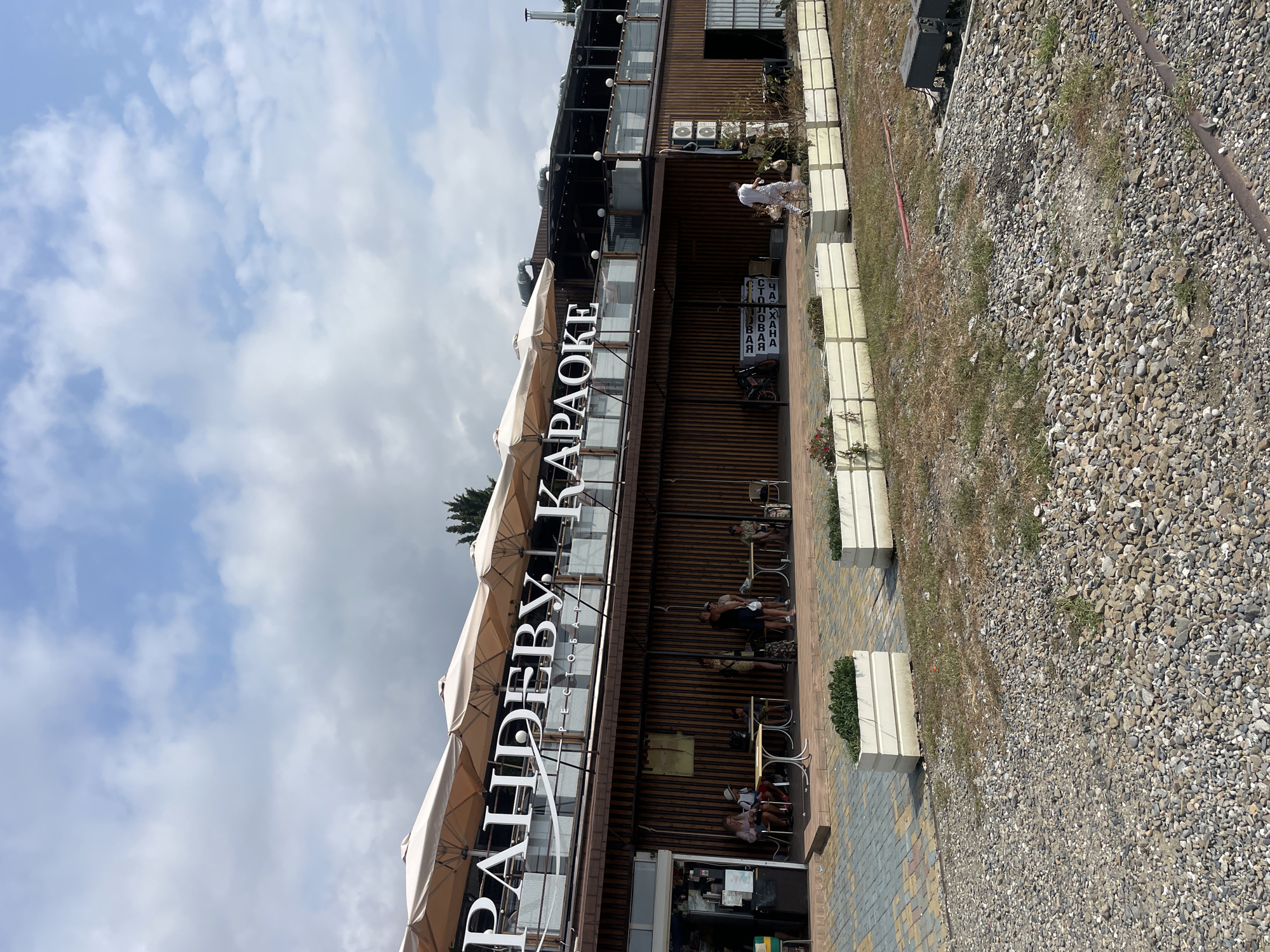 The image depicts an outdoor seating area of a karaoke bar and restaurant, identified by the large white lettering on the building that reads "РАНДЕВУ КАРАОКЕ" (RANDEVOO KARAOKE). The establishment is located in Sochi, Russia, specifically on the embankment of the Black Sea.
In the foreground, a paved walkway with a patterned design transitions to a gravelly area. Two long, rectangular white planters filled with greenery are visible along the edge of the walkway. The building itself features a two-story structure with a wooden facade on the lower level. The upper level has a modern design with glass railings and a large outdoor dining area shaded by beige umbrellas.
Several people are seated at tables outside the establishment. On the left, a group of individuals, including children, are seated at tables. In the center, a few people are standing and conversing near tables. On the right, a lone individual is seated at a table, and another person is walking away from the building towards a planter. A person in white overalls is seen walking across the gravel area in the middle ground.
Various objects are present, including tables and chairs, planters, air conditioning units mounted on the building, and a sign with Cyrillic text in a black frame. A motorcycle is parked to the left of the entrance. The sky above is partly cloudy. The overall scene suggests a casual, daytime gathering at a seaside establishment.