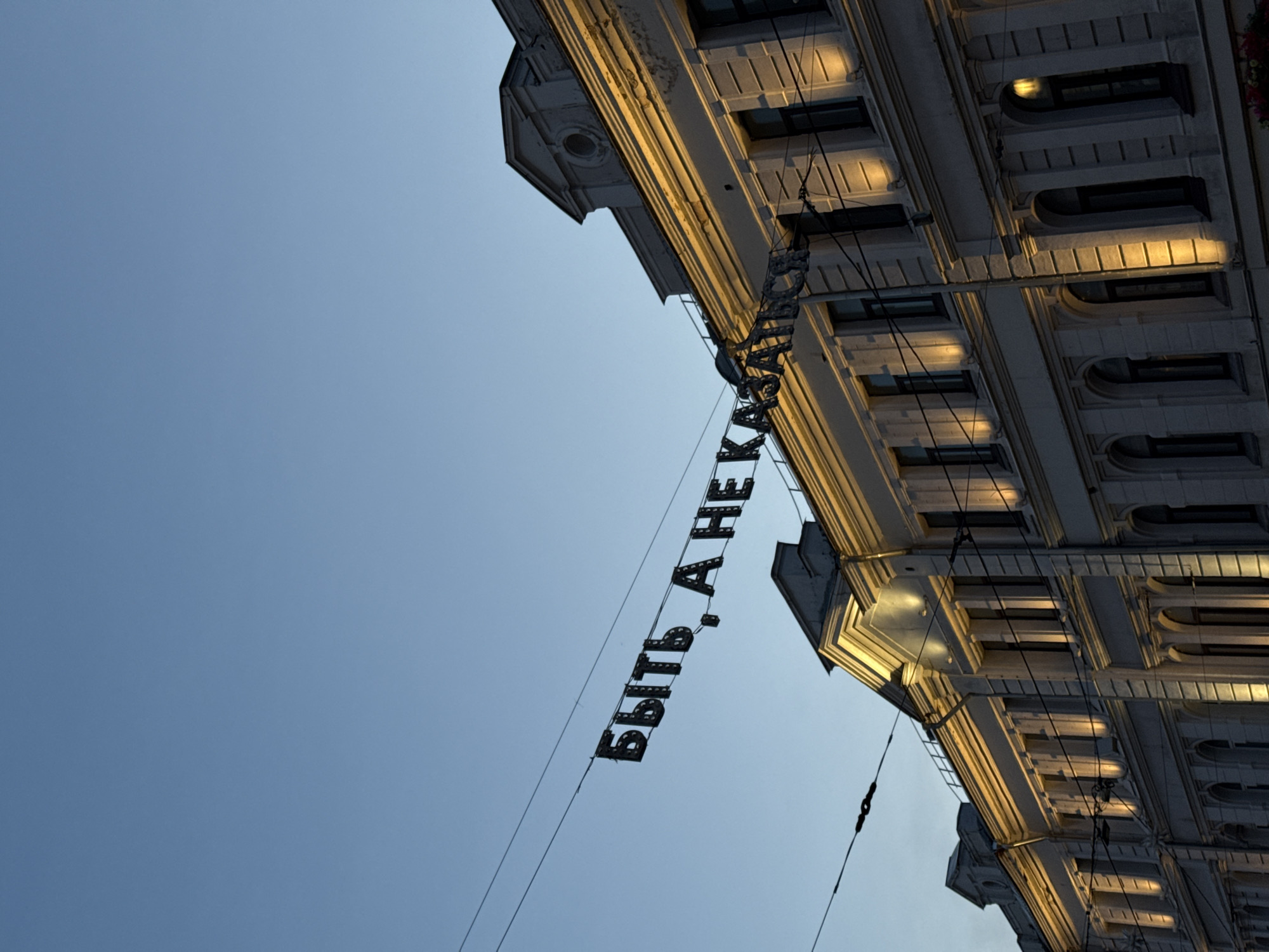 View looking up at the facade of a building in Nizhny Novgorod, Russia. The sky is light blue. A sign with Cyrillic text hangs on wires. The building is light-colored with architectural details, including windows and moldings. The windows on the building are dark, and the facade is illuminated with warm light.