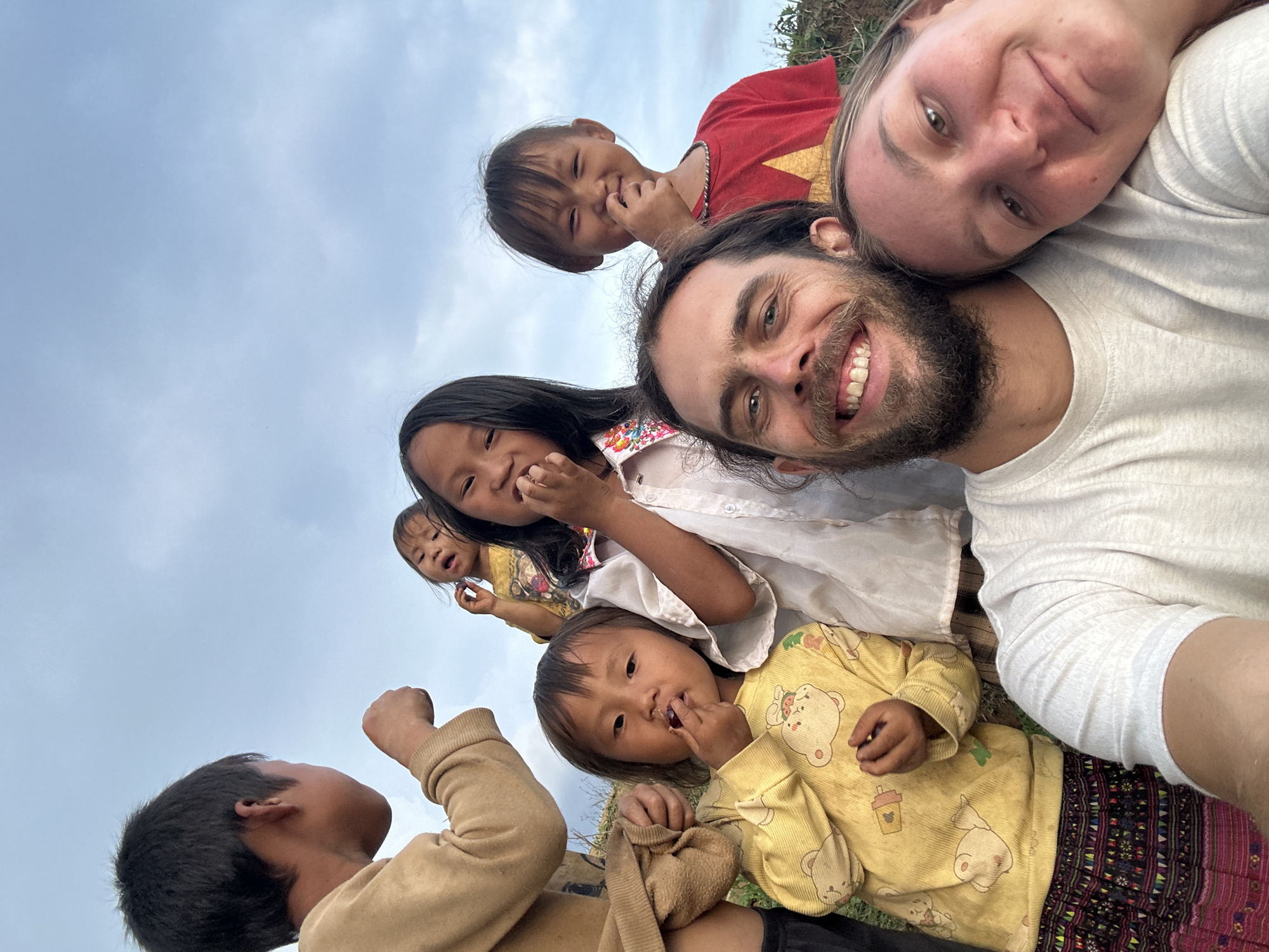 This is a warm and engaging outdoor group portrait, likely a selfie, featuring two adults and at least five children, captured from a low angle against a light blue, cloudy sky. The scene conveys happiness and close interaction.
In the foreground, an adult male with dark, bearded hair smiles broadly at the camera. Beside him, an adult female with lighter hair also smiles, her head resting near his, both wearing light-colored t-shirts. Their expressions suggest contentment and affection.
Surrounding the adults are several children of varying ages, from toddlers to older children, all looking towards the camera with curious and joyful expressions. One young child wears a vibrant red shirt adorned with a yellow star, a strong cultural symbol, and smiles with a finger near their mouth. Another girl with long dark hair, wearing a white shirt, also smiles pleasantly. A toddler in a yellow long-sleeved top patterned with bears looks directly at the camera, with a finger near their mouth. To the lower left, an older child in a light brown top is seen from behind, looking upwards towards the group.
The background is dominated by an overcast sky with soft, light blue hues and faint cloud formations, indicating daytime. A small patch of green foliage or grass is visible on the lower right, suggesting an outdoor, natural environment. The overall atmosphere is one of joyful community and friendly interaction. Given the context of Mù Cang Chải, Vietnam, the imagery, particularly the child's red shirt with a yellow star, aligns with the local setting and culture. There is no visible text in the image.