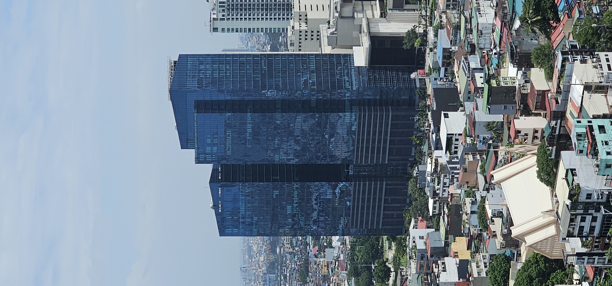The image shows an aerial view of the city of Makati, Philippines, with a focus on modern skyscrapers and densely packed residential buildings. Dominating the center are two tall, dark blue glass skyscrapers, reflecting the sky and surrounding urban landscape. To the right of these, a lighter-colored, rectangular high-rise building stands out. Below the skyscrapers, a sprawling neighborhood of smaller, multi-colored houses with various roof materials like corrugated metal and tiles is visible. A prominent, large building with a white, angular roof and spires, possibly a church or religious institution, is situated in the lower part of the frame amidst the residential structures. The overall scene depicts a contrast between contemporary urban development and older, more traditional housing in a densely populated city.