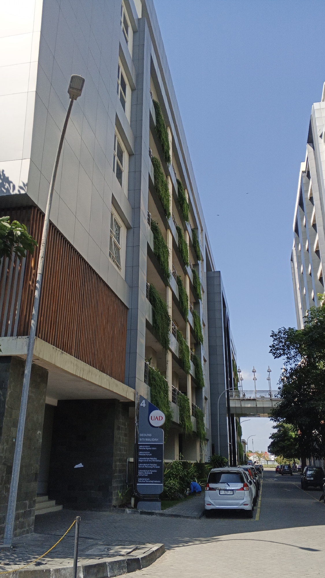 The image shows a modern building with a grey facade and numerous floors, featuring balconies with hanging green plants. A street lamp stands in the foreground to the left. A sign with the text "UAD GEDUNG SITI WALIIDAH" and a list of laboratories is visible near the entrance. Several cars are parked along the street. A pedestrian bridge connects to the building in the distance. The sky is clear and blue. The building appears to be part of a university campus in Banguntapan, Indonesia, as indicated by the signage.