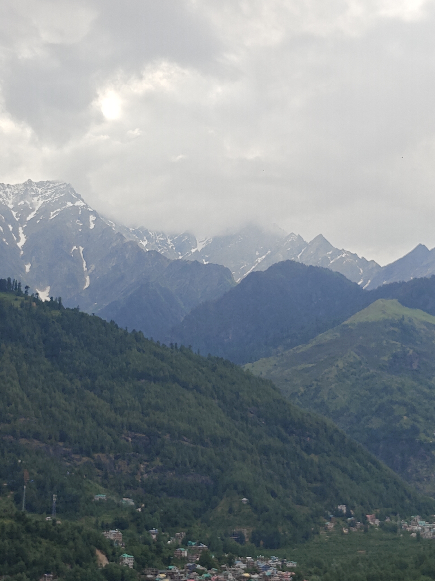 This image depicts a mountainous landscape in Vashisht, India, under an overcast sky. The upper portion of the frame is dominated by heavy grey clouds, with a bright, diffused light source visible through a break in the clouds in the upper left quadrant. The background features several layers of mountain ranges. The furthest peaks are high, rugged, and exhibit significant snow cover on their summits and upper slopes, appearing bluish-grey due to atmospheric haze. Closer mountain ranges, also snow-dusted, rise behind and blend into densely forested, dark blue-green slopes in the mid-ground. The immediate foreground and mid-ground are characterized by steep, lush green mountainsides covered in dense deciduous or coniferous forest.
Nestled within the lower slopes and valleys of these green mountains, particularly in the lower half of the image, is a human settlement identified as Vashisht. Numerous multi-story buildings with varied roof colors, including blue, green, grey, and orange, are visible, clustered together, indicating a populated area. Modern infrastructure is present, with at least two tall, slender communication towers (likely cellular towers) erected on a forested slope in the lower-left quadrant. No specific individuals, animals, or explicit human actions are discernible within the frame due to the distance and scale. The overall lighting is soft and diffused, characteristic of an overcast day.