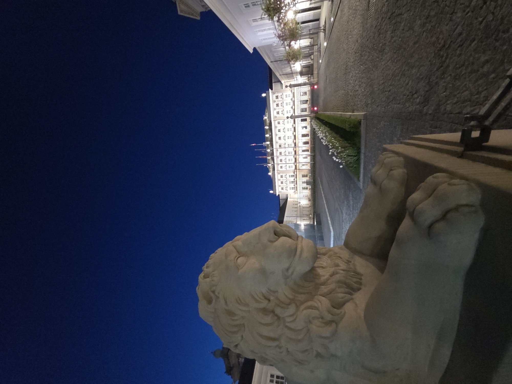 A long-shot, outdoor, nighttime view shows a stone lion statue facing toward a building in Warsaw, Poland. The lion statue is composed of beige stone and has two paws in the foreground on the right side of the image. In the background is a building with a long row of windows, flags, and lights. A grey stone walkway is between the lion statue and the building. A strip of green grass and white flowers separates the walkway from the building. The sky above is a dark blue.