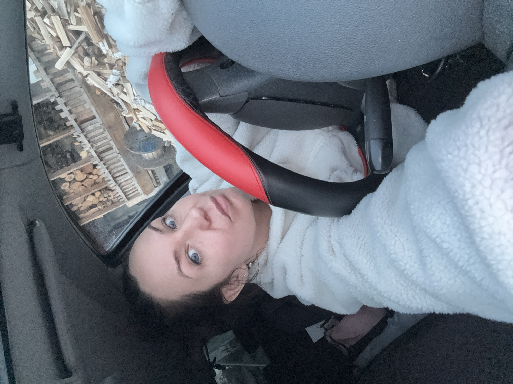 The image is a vertical, close-up shot taken from inside a car in Chaikovsky, Russia. The primary subject is a woman with striking blue eyes and dark hair, looking directly at the camera with a neutral expression. She is wearing a white, fluffy long-sleeved garment, possibly a fleece or bathrobe, and has a small earring visible on her left ear.
The interior of the car is partially visible, including a red and black steering wheel cover in the upper right, and portions of the grey dashboard and seat. Through the car's window on the left, an outdoor scene is depicted. This background features a large stack of neatly arranged firewood, predominantly light-colored wood like birch, piled against wooden support structures. A dark, possibly wooden, industrial-style spool or cable reel is also seen amidst the firewood.
The image appears to be taken during the daytime, with soft, natural lighting suggesting an overcast sky. The presence of significant firewood stacks indicates a cold climate or preparedness for winter. There is no visible text in the image. The woman's direct gaze implies she is aware of being photographed, possibly a selfie.