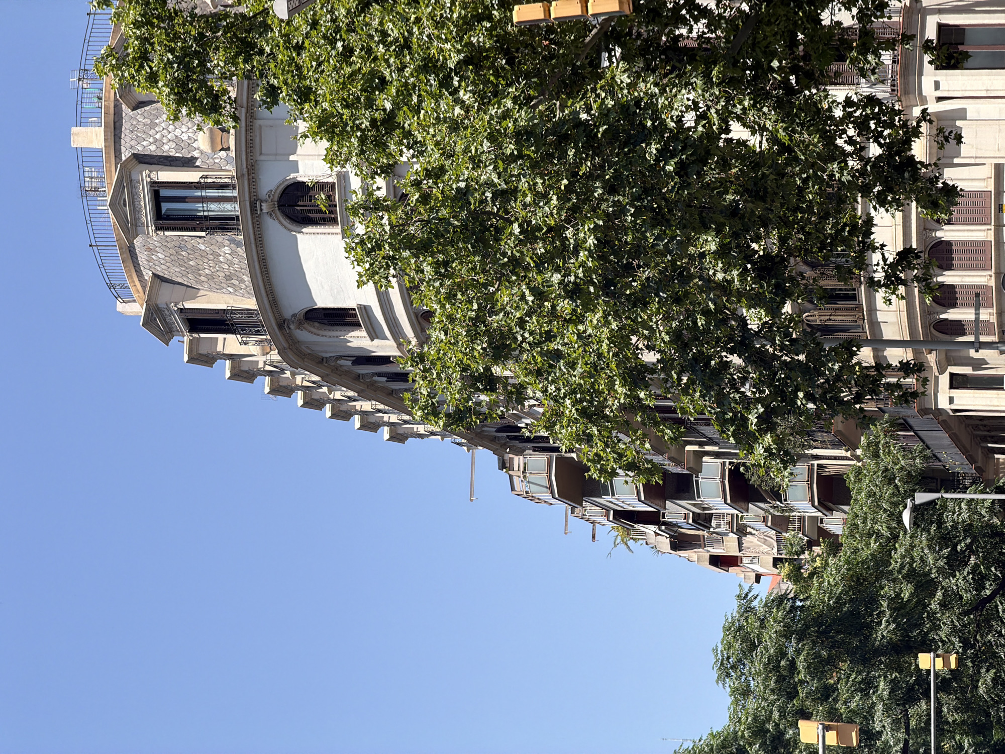The image shows a street view in Barcelona, Spain, on a clear, sunny day. A tall, ornate building with a mansard roof and multiple balconies dominates the right side of the frame. Green trees with dense foliage partially obscure the building's facade. In the foreground, to the left, a traffic light with three yellow signals is visible. The sky is a uniform, bright blue.