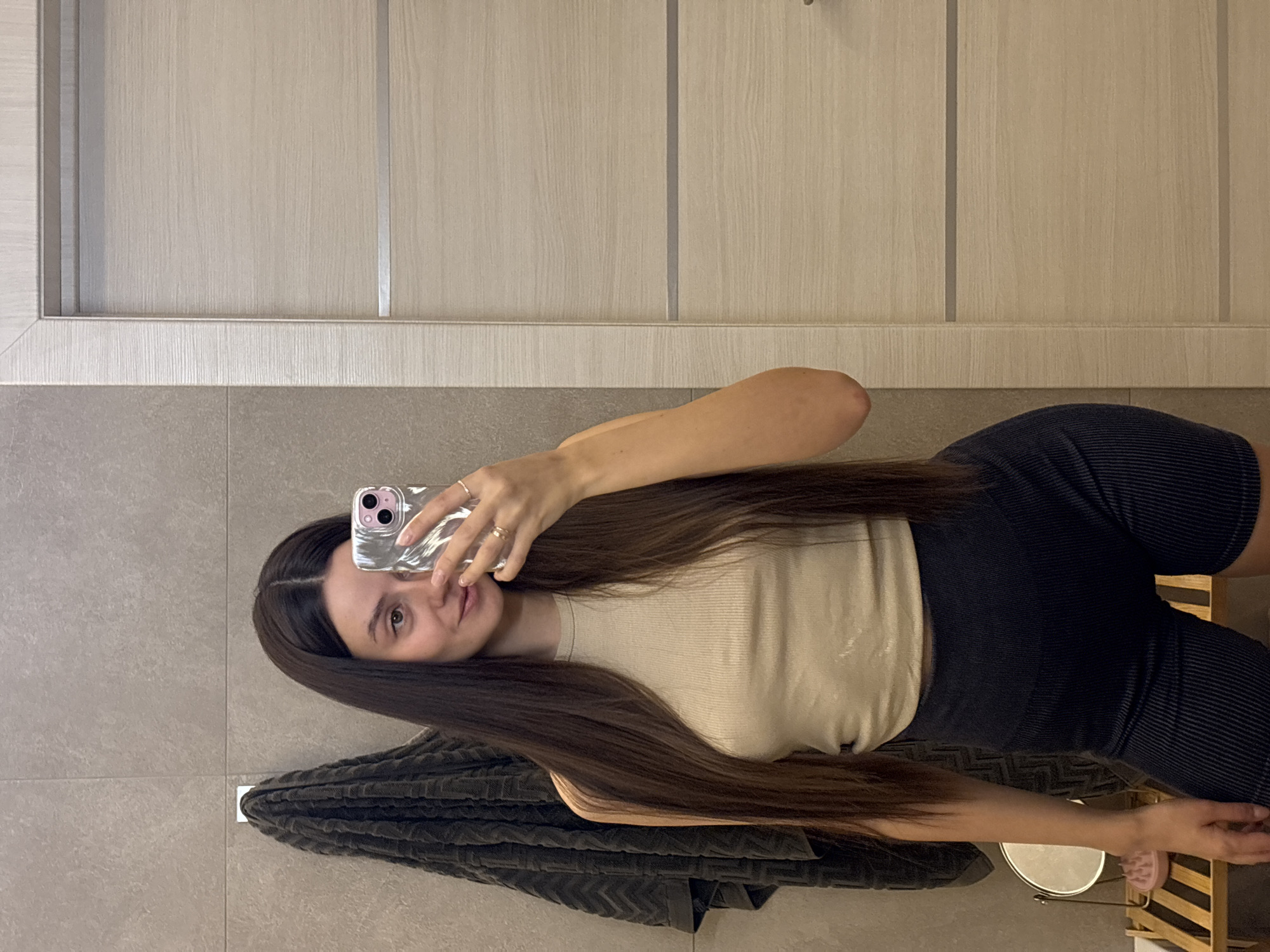 The image, presented in a sideways orientation, captures a young woman taking a mirror selfie. She has long, straight dark brown hair, parted down the middle, which extends well past her waist. She wears a light beige, ribbed sleeveless tank top and dark blue or black ribbed shorts. Her left hand, adorned with rings on the index and ring fingers, holds a smartphone with a clear case, reflecting a triple camera lens setup. She looks towards the camera/mirror with a calm expression and a slight smile.
The setting appears to be an indoor room, likely a bathroom or dressing area, characterized by light gray tiled walls and floor. A dark gray, textured towel hangs on a horizontal rail behind her. Above the tiled section, in the background, are light-colored wooden cabinets or wardrobe doors featuring vertical silver-colored strips, suggesting a modern design. Towards the bottom right of the image, a wooden stool or small shelf is partially visible, along with a small, round standing mirror and what appears to be a pink slipper or sandal. The ambient indoor lighting makes the specific time of day and outdoor weather indistinguishable. The scene is located in Krasnoyarsk, Russia. No visible text is present in the image.