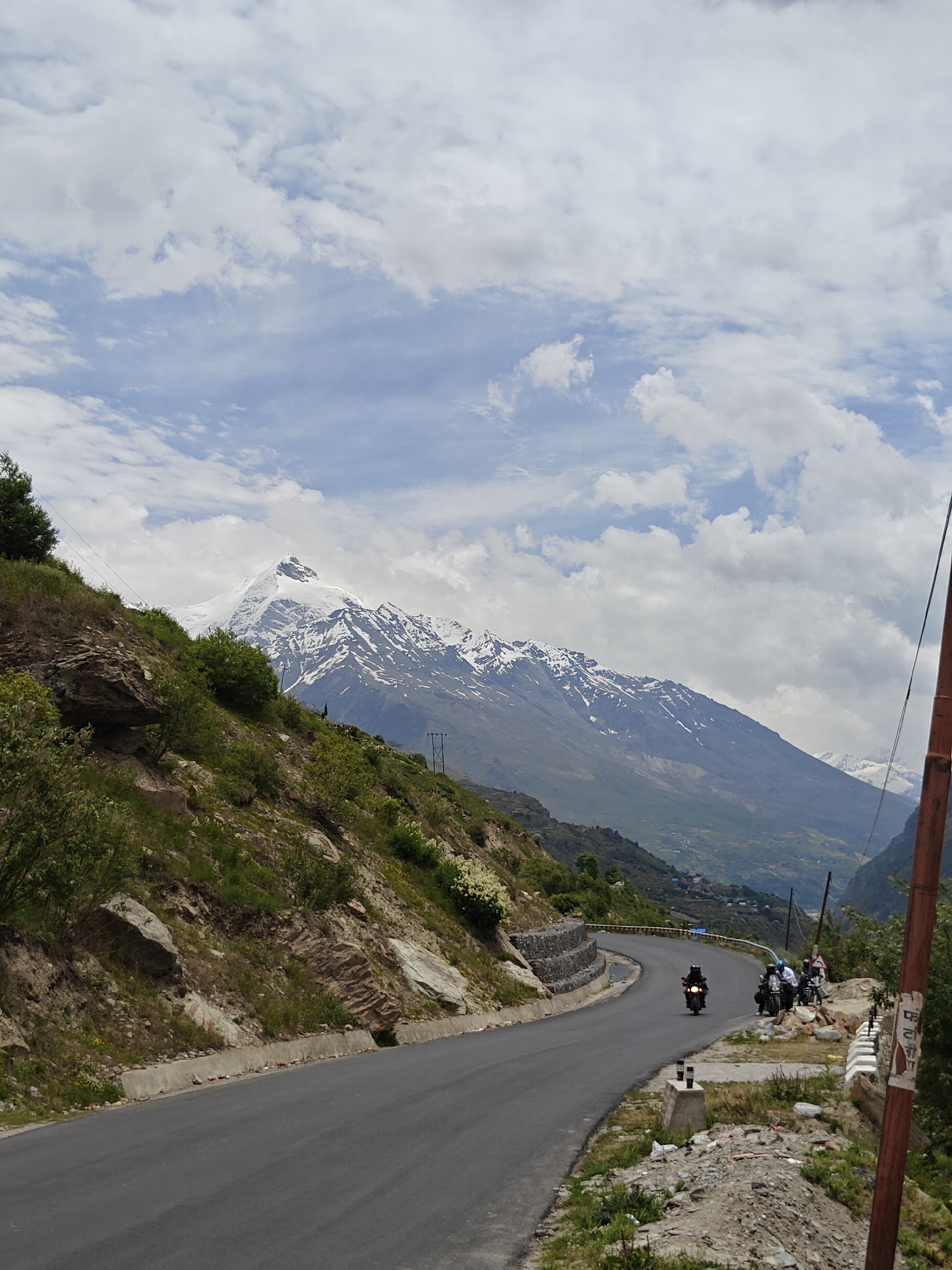 The image depicts a mountainous landscape with a winding road. In the foreground, the road curves to the left, bordered by a grassy slope with rocks. Further back, the road continues, with a group of motorcyclists riding on it. The motorcycles appear to be traveling uphill.
The background features a majestic snow-capped mountain range under a cloudy sky. The slopes of the mountains are covered in greenery, with some scattered houses visible in the distance, suggesting a rural or semi-rural settlement. The overall scene suggests a scenic route in a mountainous region, possibly in India, given the mention of Jagle.
The road appears to be paved and well-maintained, suitable for vehicular traffic. The presence of power lines on poles indicates infrastructure development in the area. The lighting suggests it is daytime, with diffused sunlight filtering through the clouds. The image captures a sense of travel and adventure in a natural, rugged environment.