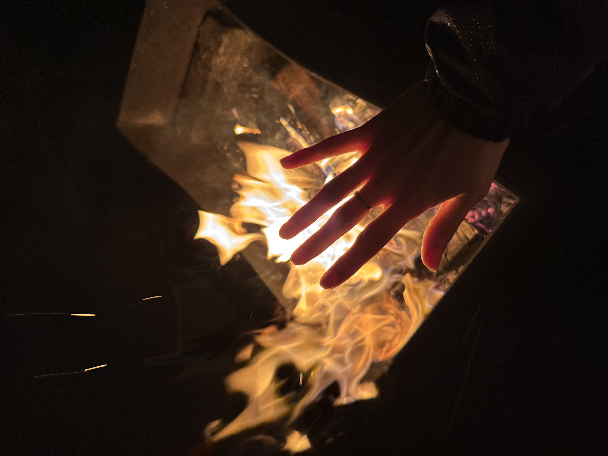 A hand with a ring on its ring finger is extended over a fire burning in a metal container. The fire is the primary light source, illuminating the hand and the immediate surroundings. A dark-colored sleeve is visible in the upper right. The environment is mostly dark, suggesting an outdoor setting at night. The image does not contain sufficient information to verify the location in Mayak, Russia.