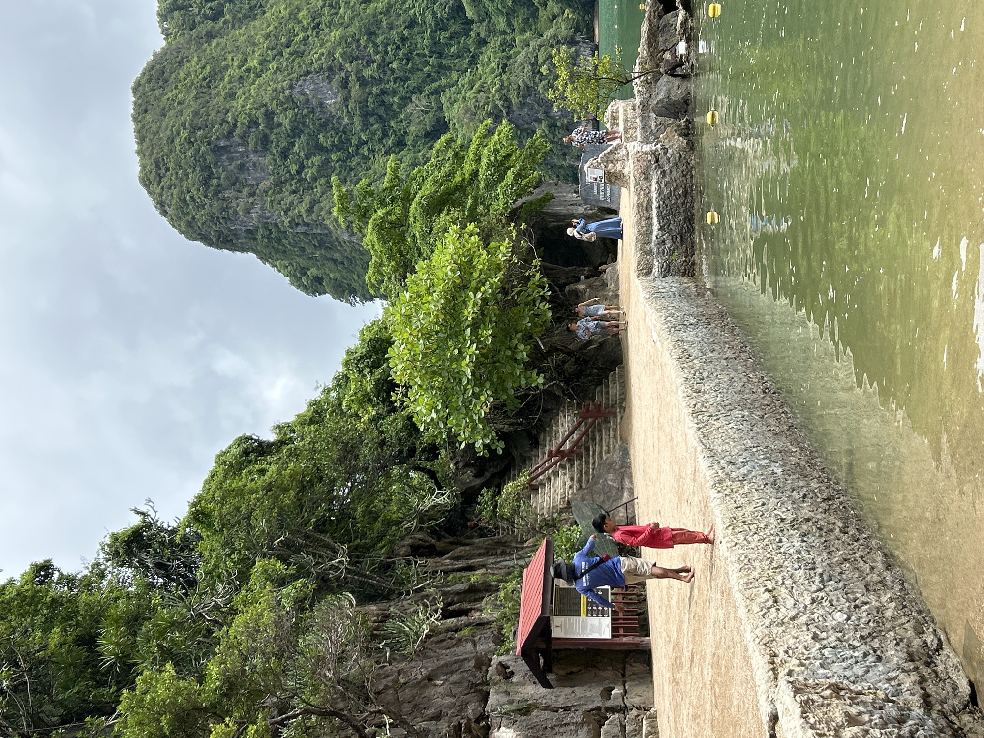 An outdoor shot captures a tropical island scene in Tambon Krasom, Thailand, with a focus on the shoreline and a forested mountainside. The foreground is dominated by clear, shallow green water lapping against a low stone wall. A sandy path runs along the wall, where several people are visible. To the left, a man in a blue shirt and shorts, wearing a hat, gestures towards a young child in a red outfit. Behind them, a small red-roofed structure with informational posters stands near a rocky cliff face. Further back, a set of stone stairs ascends towards the lush, green vegetation that covers the steep mountain in the background. Several other individuals are scattered along the path and near the stairs, some appearing to be tourists taking in the scenery. The sky is overcast, suggesting a cloudy day.