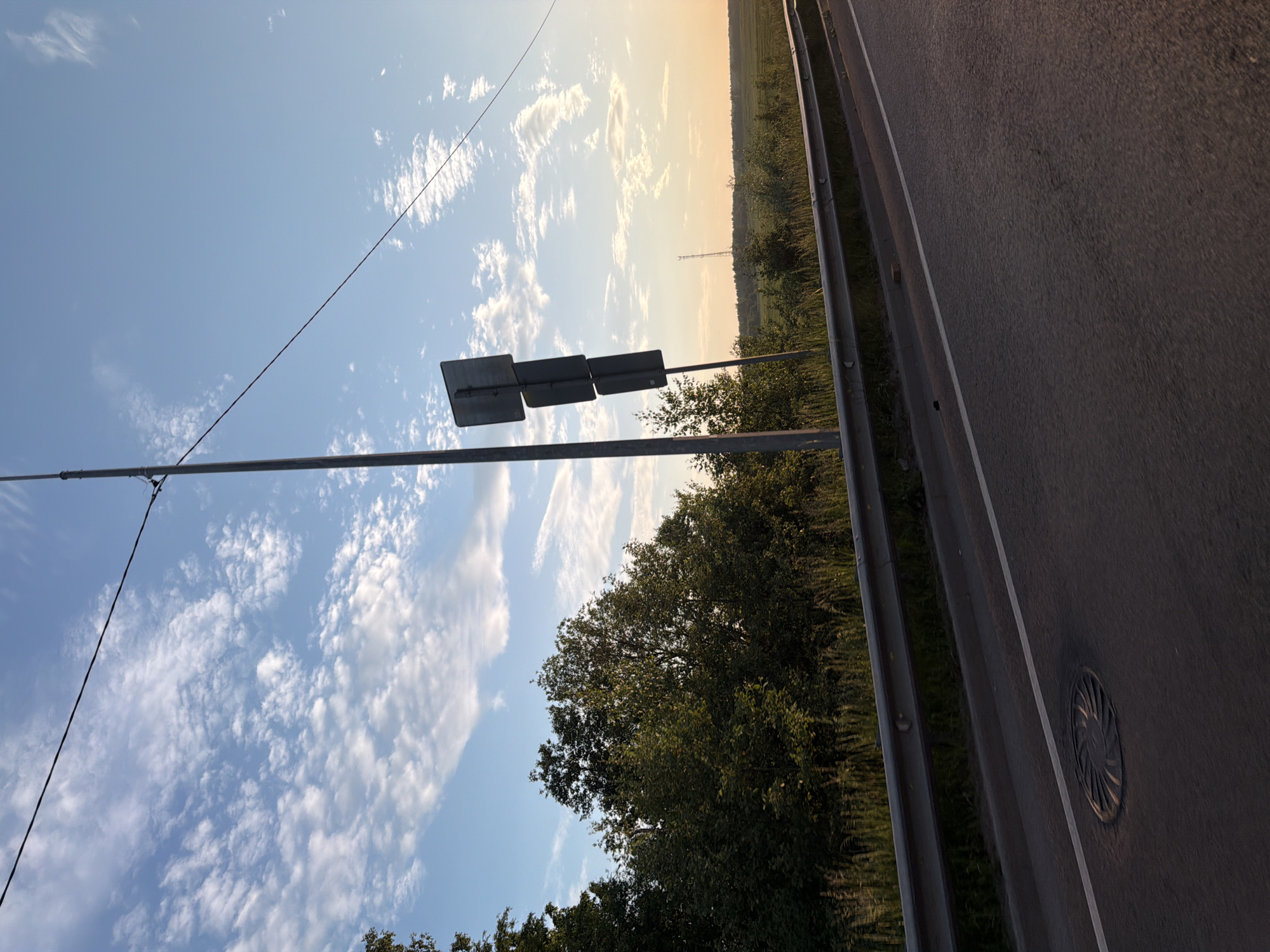 The image depicts a roadside scene under a cloudy blue sky at sunset. A tall metal pole with three stacked square signs is positioned on the left side of the frame, near a dense line of green trees. The road, paved and with a white line marking, occupies the foreground and extends to the right. A metal guardrail runs parallel to the road, separating it from a grassy area and the trees. In the distance, a vast green field is visible, stretching towards a soft orange horizon. A solitary cell tower stands in the middle ground, silhouetted against the sky. The overall atmosphere is serene, capturing the quiet beauty of a rural landscape at dusk.