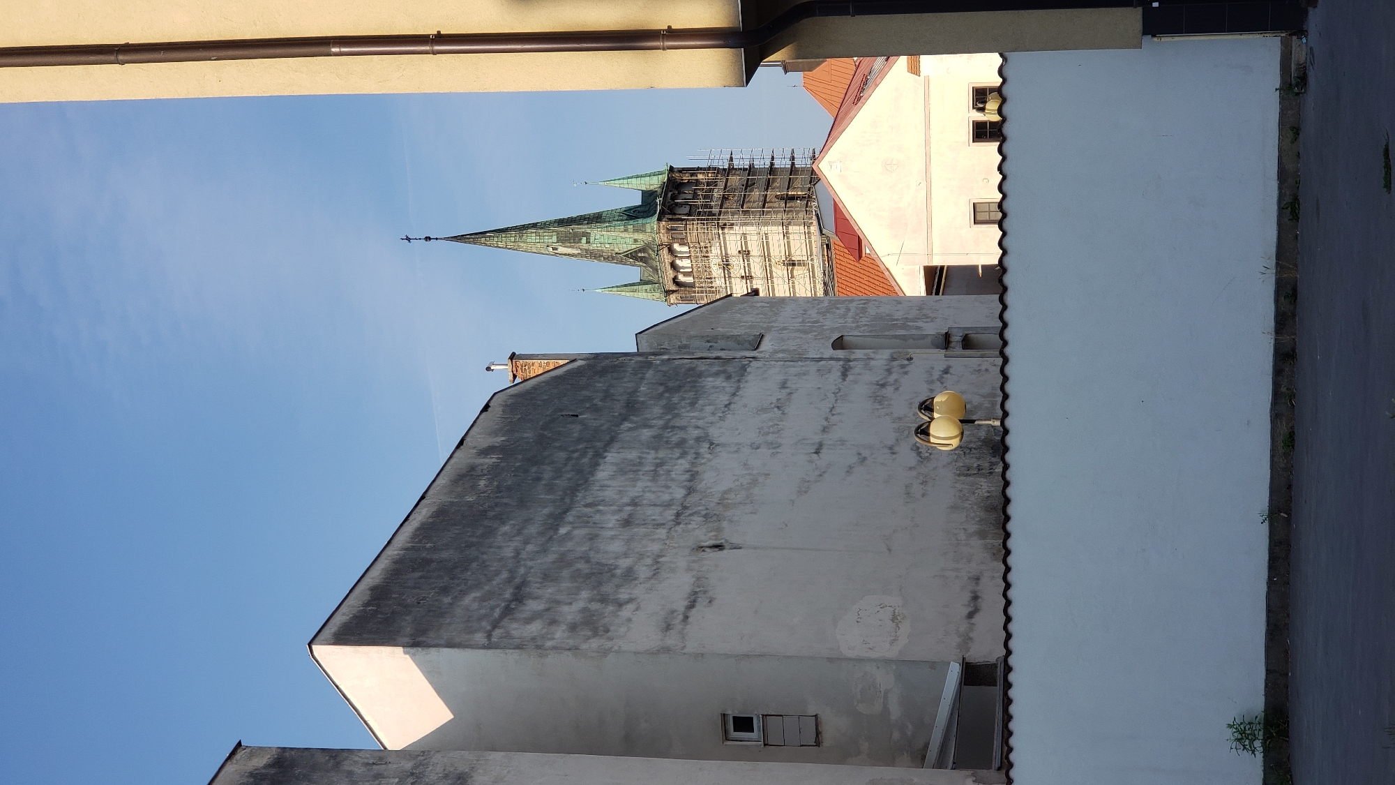 The image shows a street view with buildings and a church tower under a clear blue sky. The foreground is dominated by a white wall with a dark, wavy trim. A paved area is visible in front of the wall, with a few small plants growing along the base. Behind the wall, two yellow street lamps are mounted on a pole.
To the left, a tall, grey building with a weathered facade takes up a significant portion of the frame. It has a small, white-framed window. Adjacent to this building, another grey structure is partially visible.
In the background, the spire of a church, partially covered by scaffolding, rises above other buildings. The spire is green and has a cross at its apex. Other buildings with red-tiled roofs and light-colored walls are visible between the grey buildings and the church. The overall scene suggests an urban environment, likely in a European city.
Based on the architectural style and the presence of the prominent church tower, this scene is likely located in the city of Prešov, Slovakia, specifically in the vicinity of the St. Nicholas Cathedral (also known as the Roman Catholic Cathedral of Saint Nicholas), which features a similar spire and is undergoing renovation, as indicated by the scaffolding. The composition, with its narrow street view and looming architecture, is typical of older European city centers.
