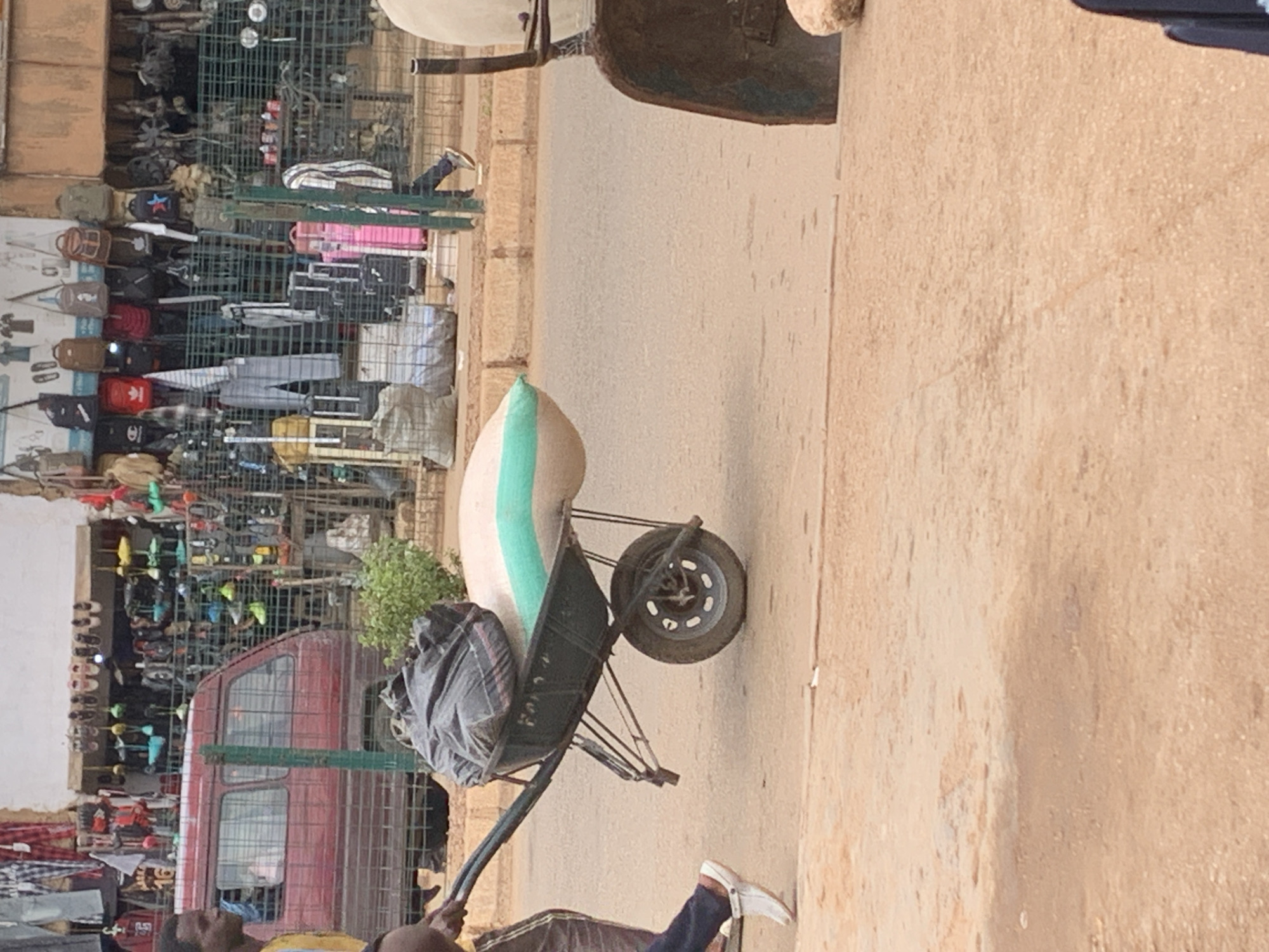 A person is pushing a wheelbarrow filled with goods down a street, likely in a market. The scene is outdoors, and the lighting suggests it is daytime. The ground appears to be a paved road or pathway. The background shows a market stall with various items on display, including clothing and accessories, organized on metal shelving units. A red van is also visible parked behind the stall. The wheelbarrow contains a large, light-colored sack with a green stripe and a dark-colored bundle wrapped in plastic. The person pushing the wheelbarrow is wearing dark pants and white shoes. The overall impression is of a busy marketplace or street scene in Nigeria, specifically Bukuru. The weather appears to be clear and sunny, contributing to the bright illumination of the scene. The image captures a moment of daily commerce and activity in a bustling environment.
